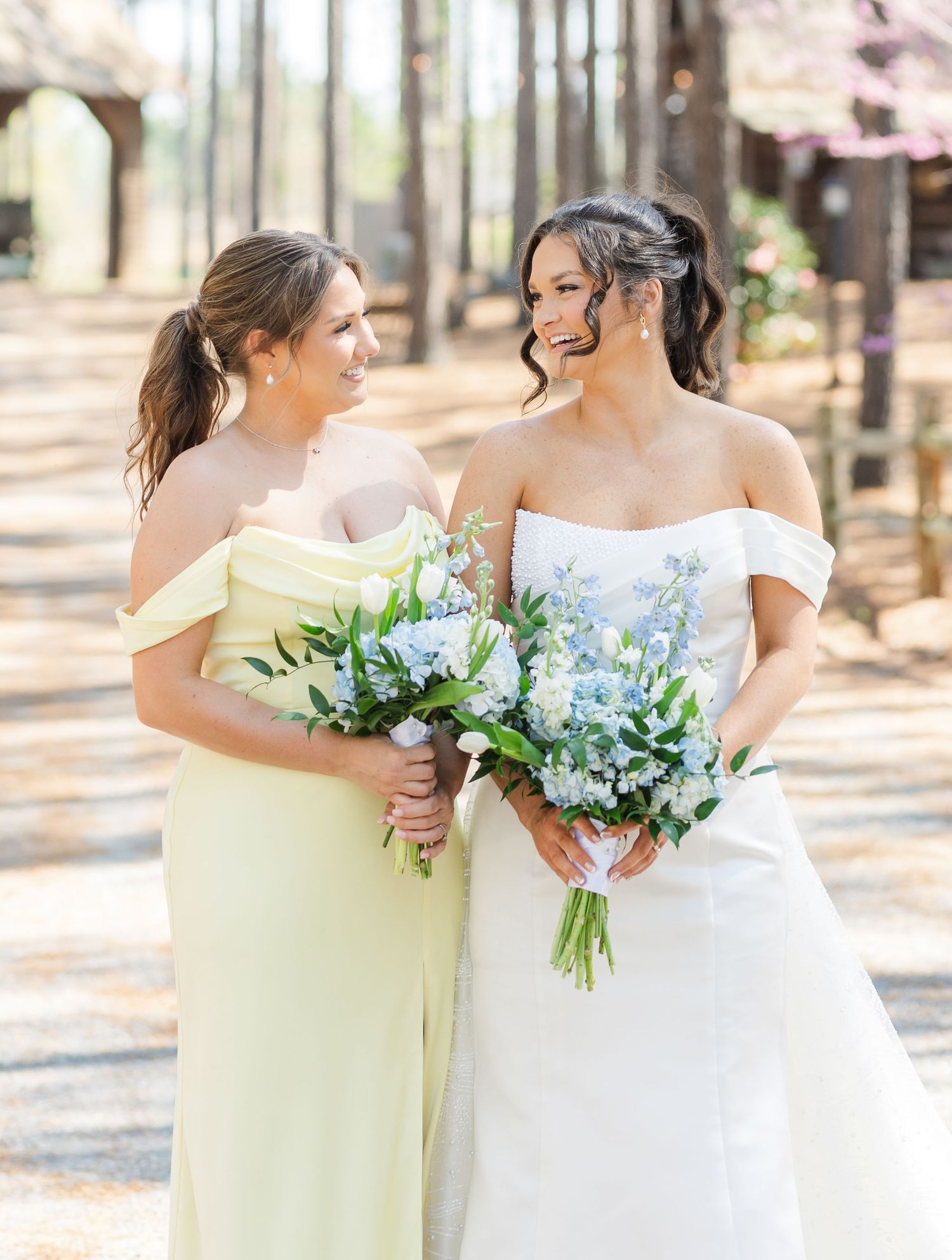 Bride in off-shoulder gown and bridesmaid in yellow dress holding bouquets of blue and white flowers.