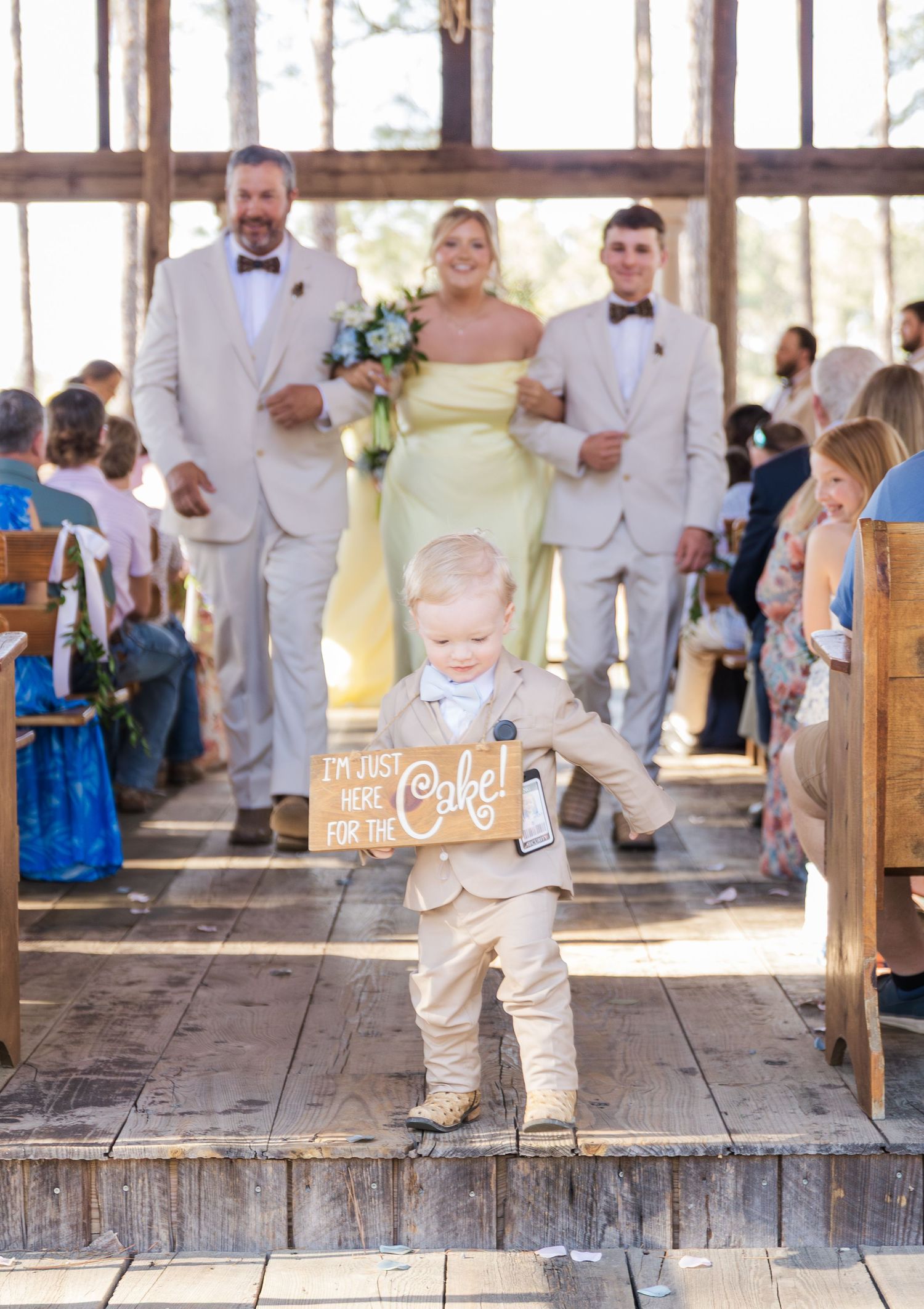 Ring bearer carries humorous cake sign down aisle as wedding party follows in rustic venue.