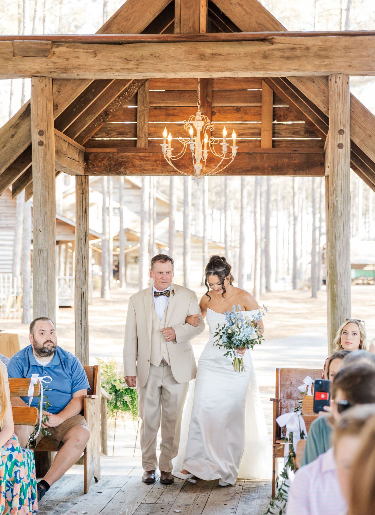 Bride walking down outdoor aisle with escort under wooden pergola with chandelier at rustic wedding venue.