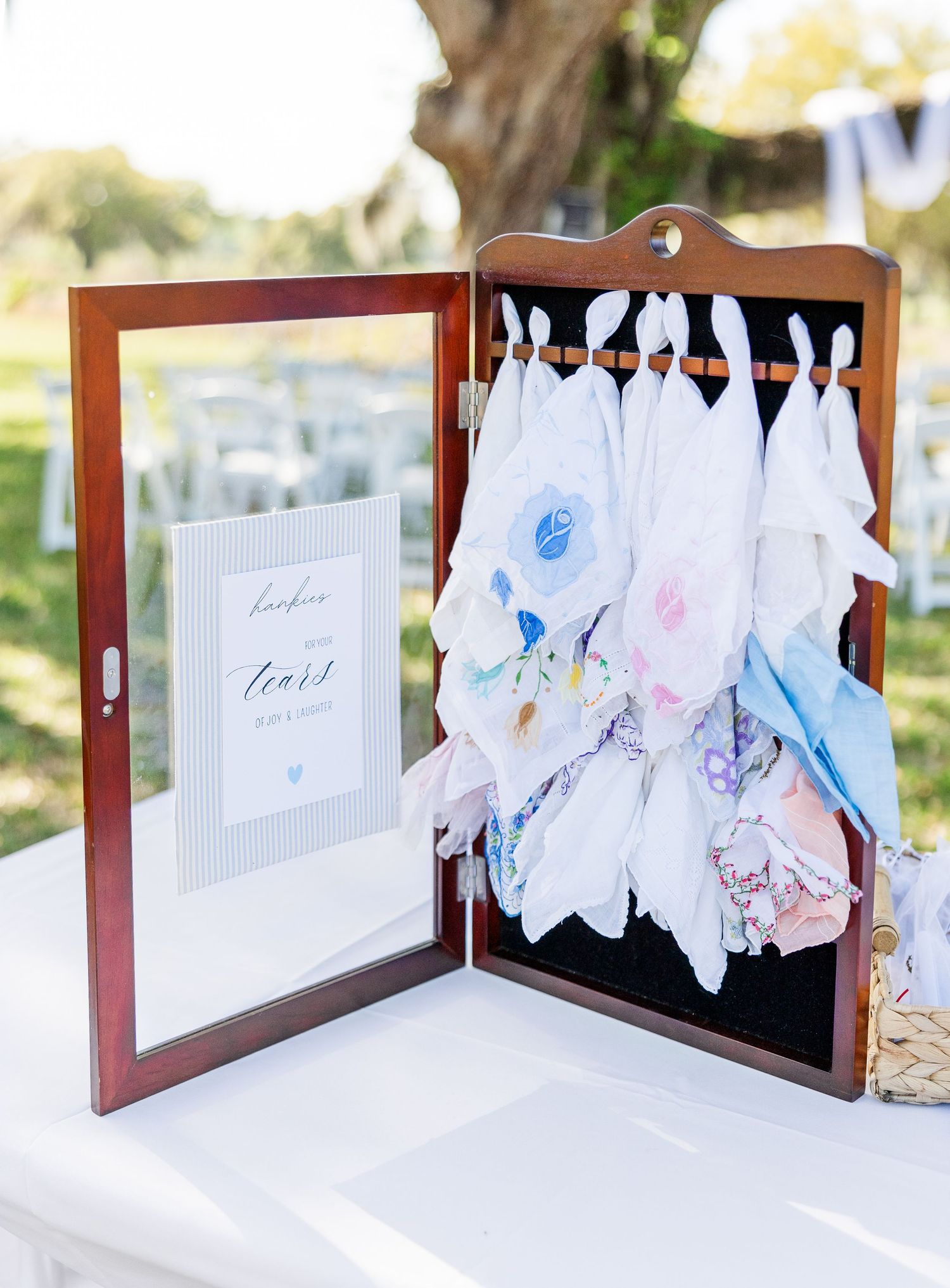 Wooden display case holding colorful handkerchief favors for wedding guests beside a framed sign outdoors.