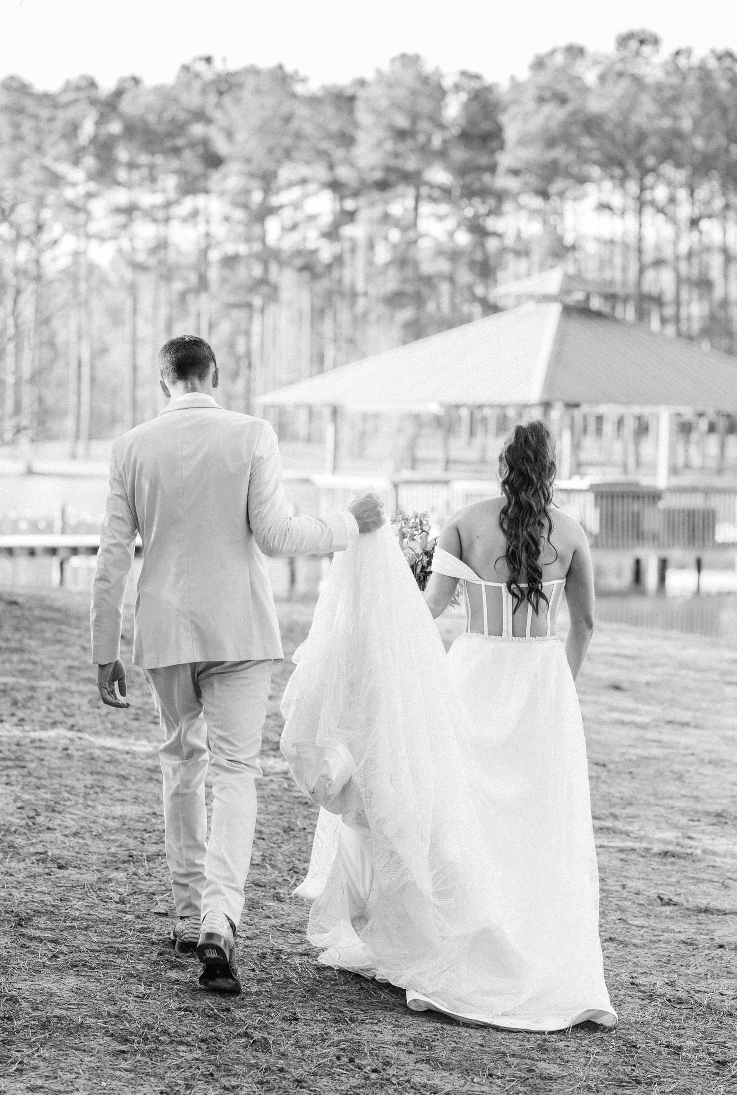 Black and white photo of bride and groom walking hand in hand toward a lakeside gazebo on their wedding day.