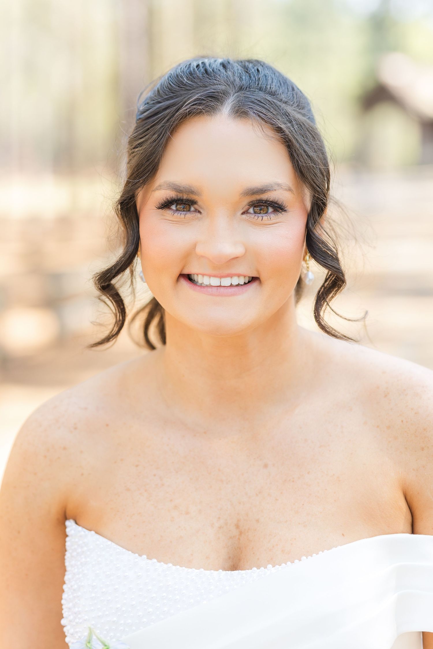 Close-up portrait of a smiling bride in a strapless white gown in a pine forest setting.