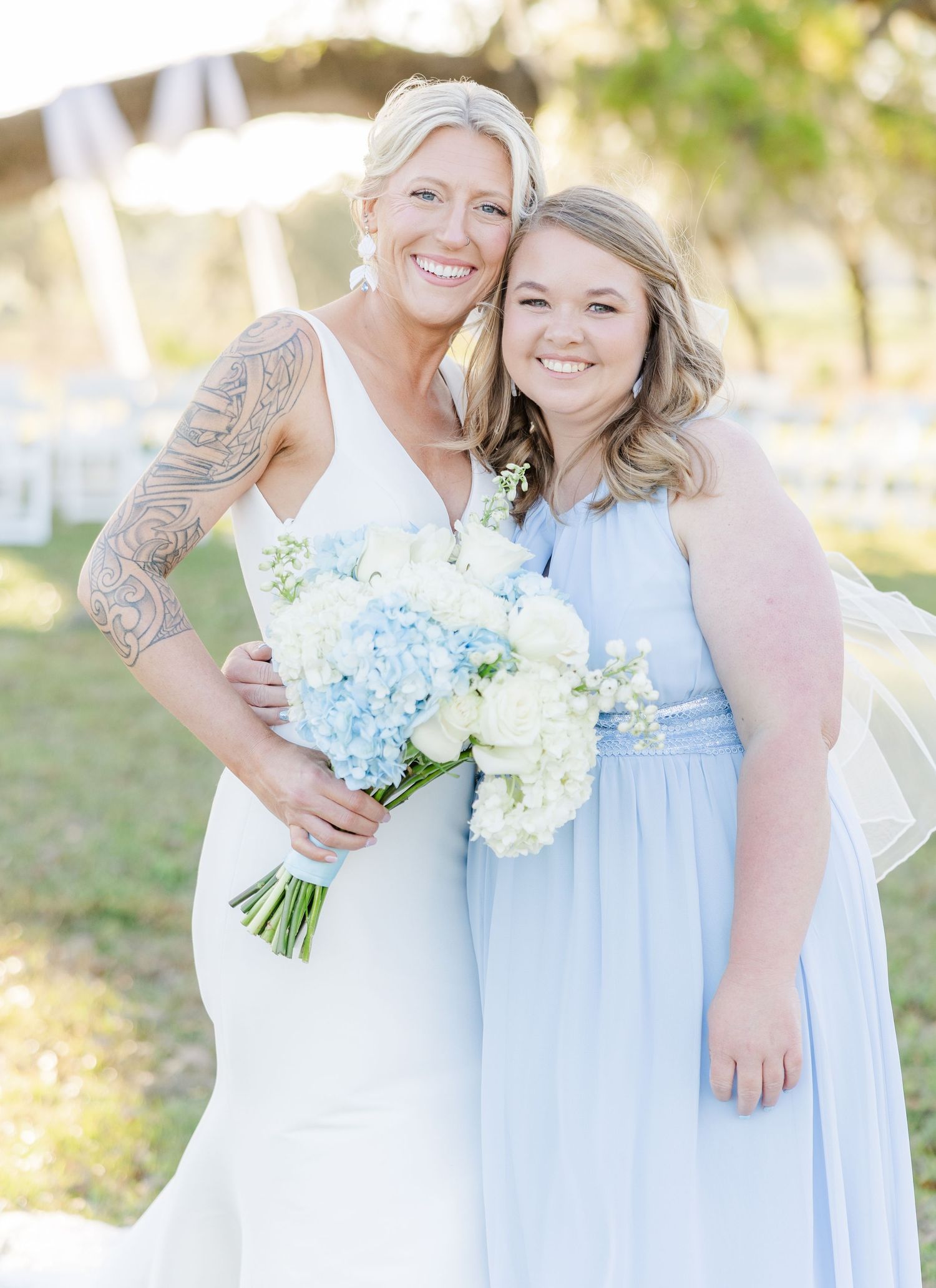 Bride with sleeve tattoo and bridesmaid in light blue dress pose together holding white and blue hydrangea bouquet.