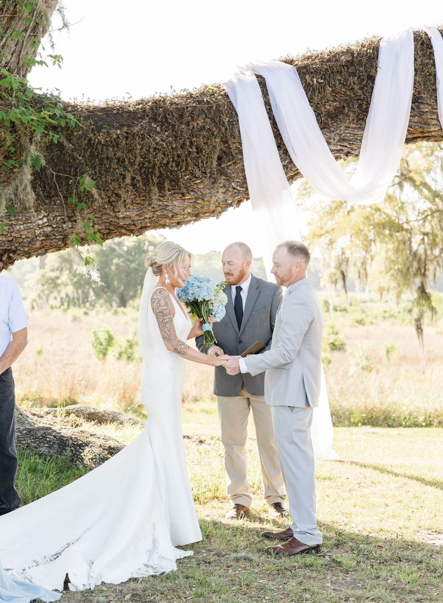 Bride and groom exchange vows under a rustic moss-draped arch with white fabric at an outdoor ceremony.
