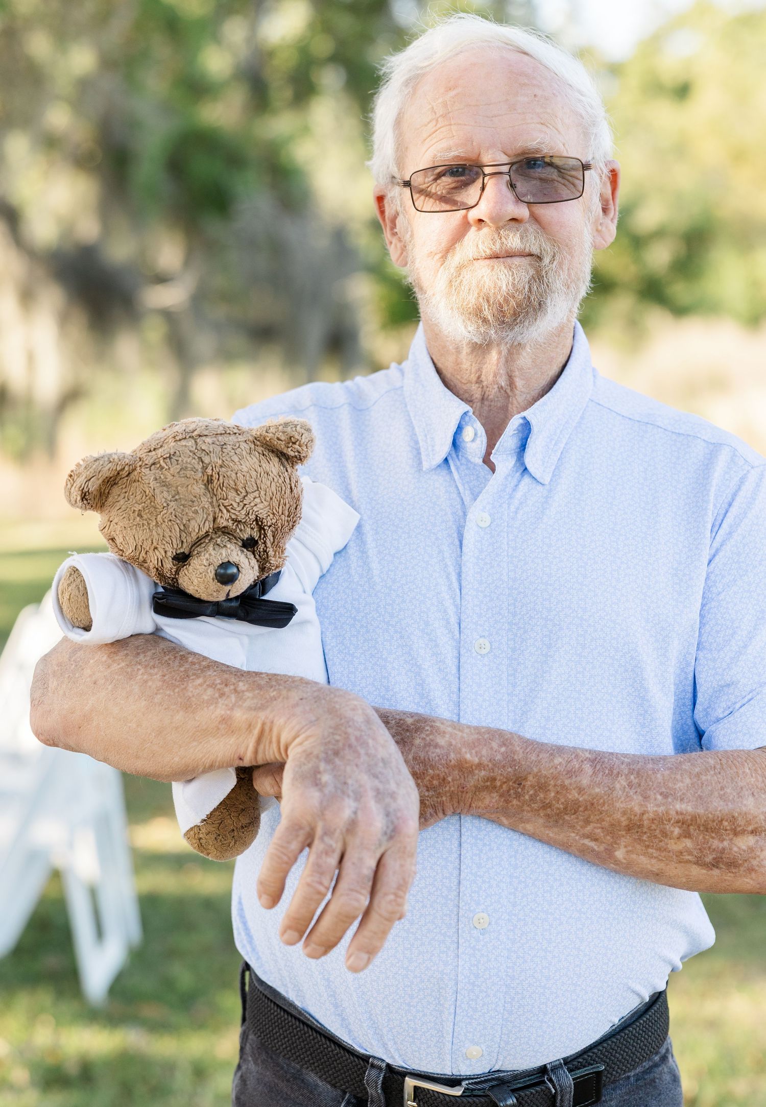 Elderly man with white beard holds a brown teddy bear with bow tie outdoors at a wedding venue.