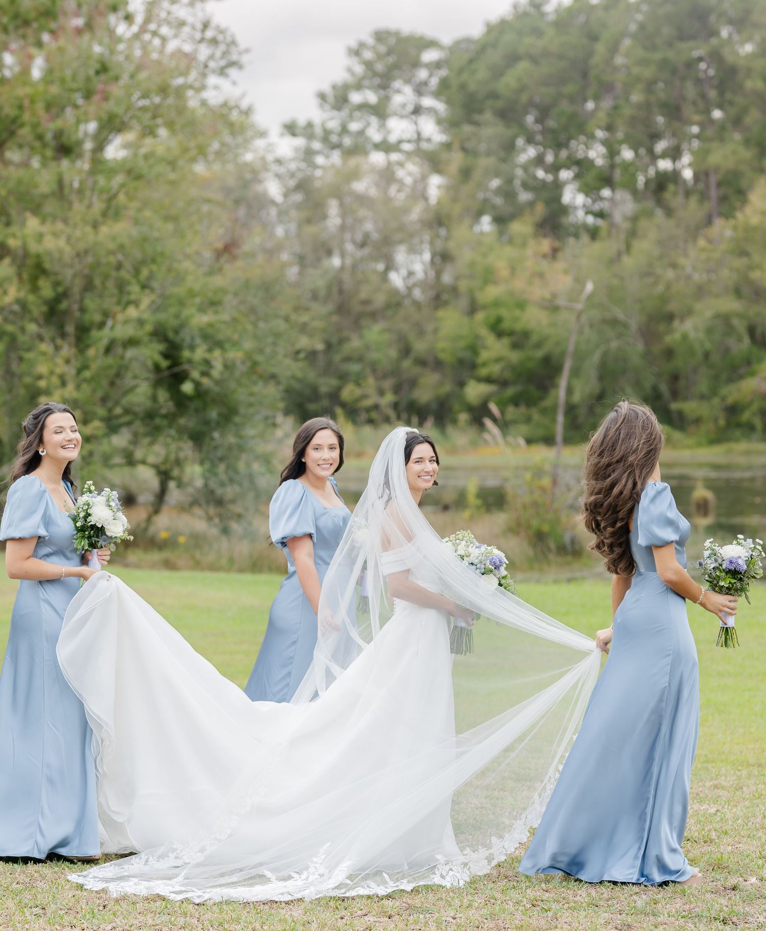Wedding party in light blue bridesmaid dresses poses with bride in flowing white gown outdoors on grassy field.
