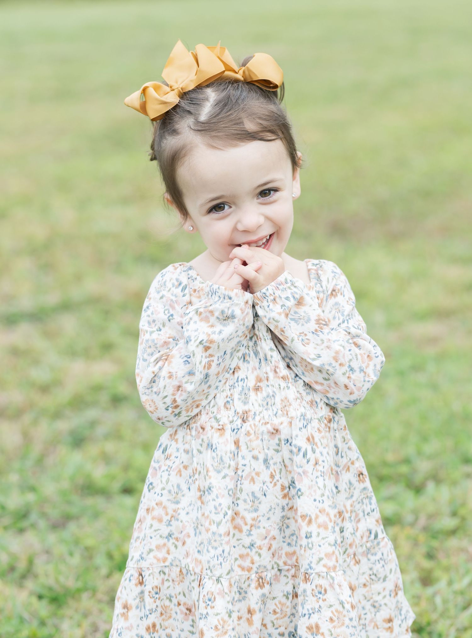 Sweet toddler in floral dress and yellow bow playing in a grassy outdoor setting.