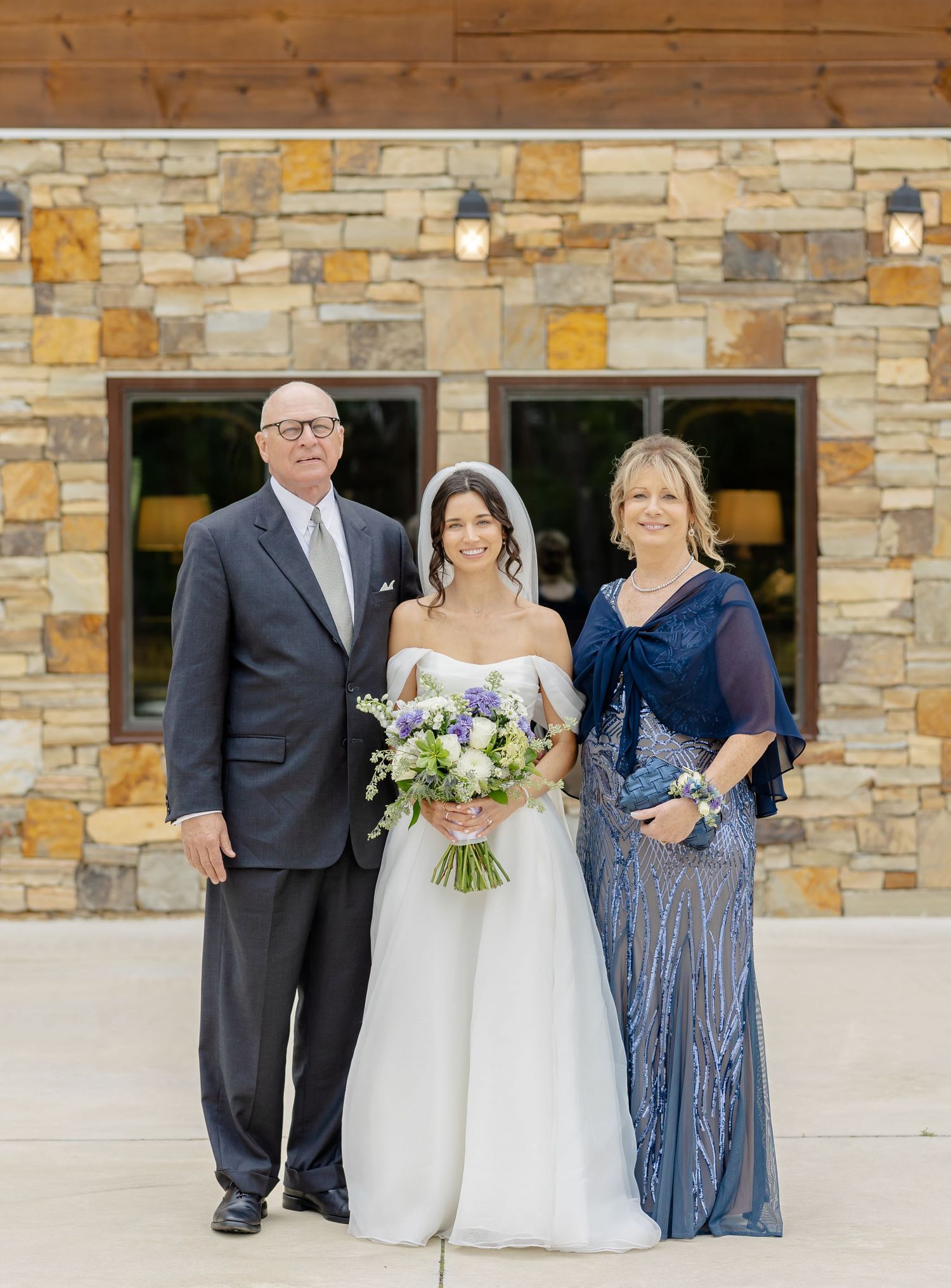 Wedding party poses in front of stone building with bride holding purple and white bouquet.