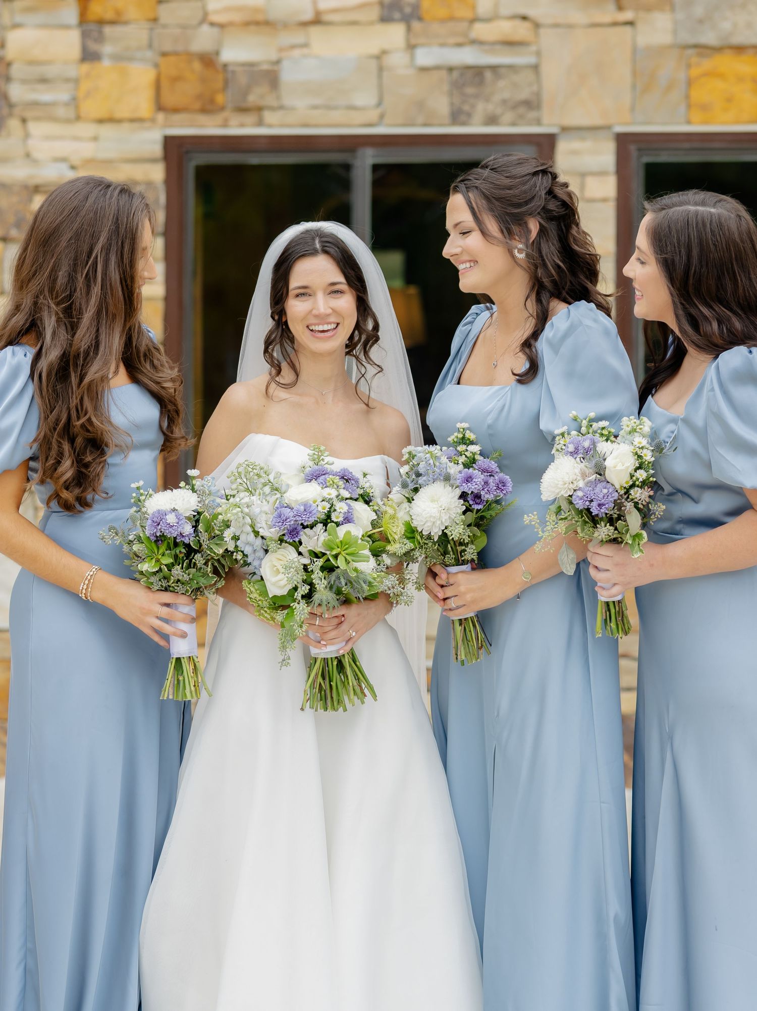 Bridesmaids in light blue dresses hold white and purple bouquets while standing together at a wedding.