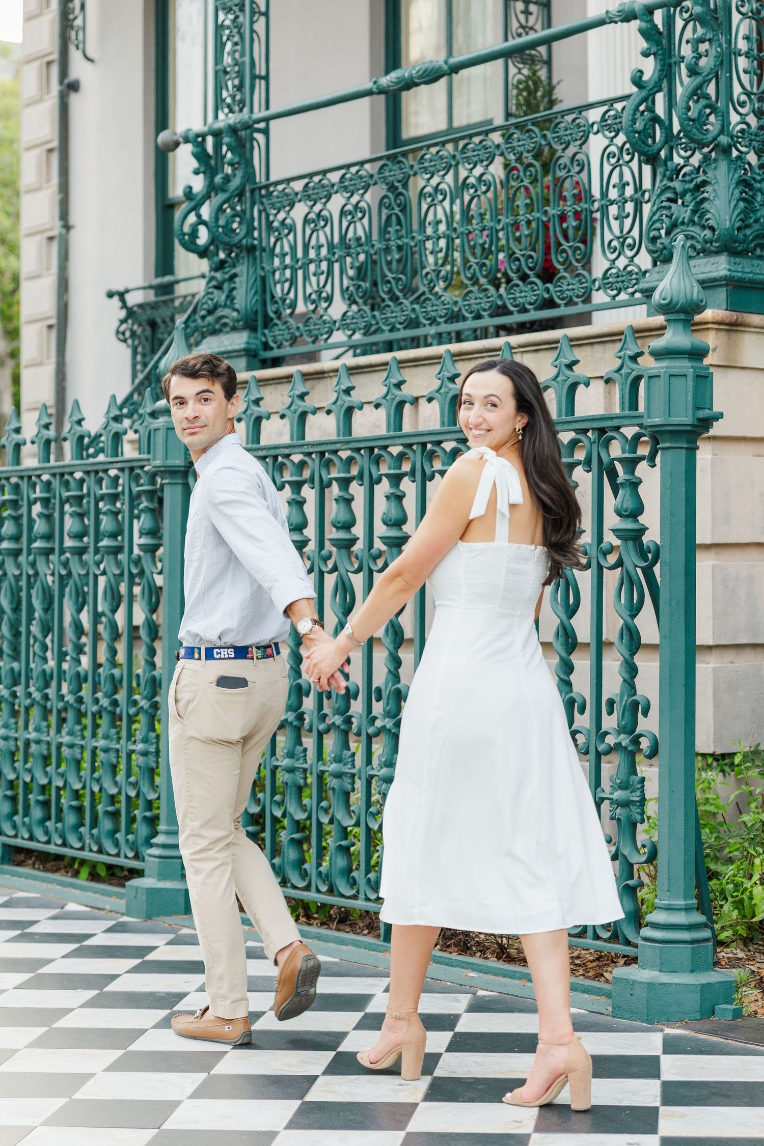Couple holds hands and smiles while posing by an ornate green iron fence on a checkered sidewalk.