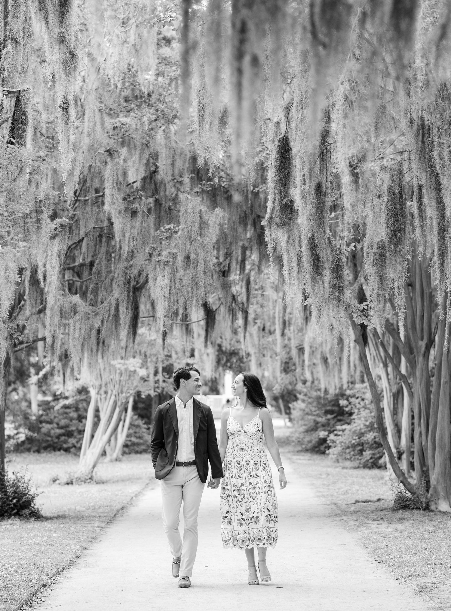 A couple walks hand-in-hand along a tree-lined path draped with Spanish moss in a black and white photo.