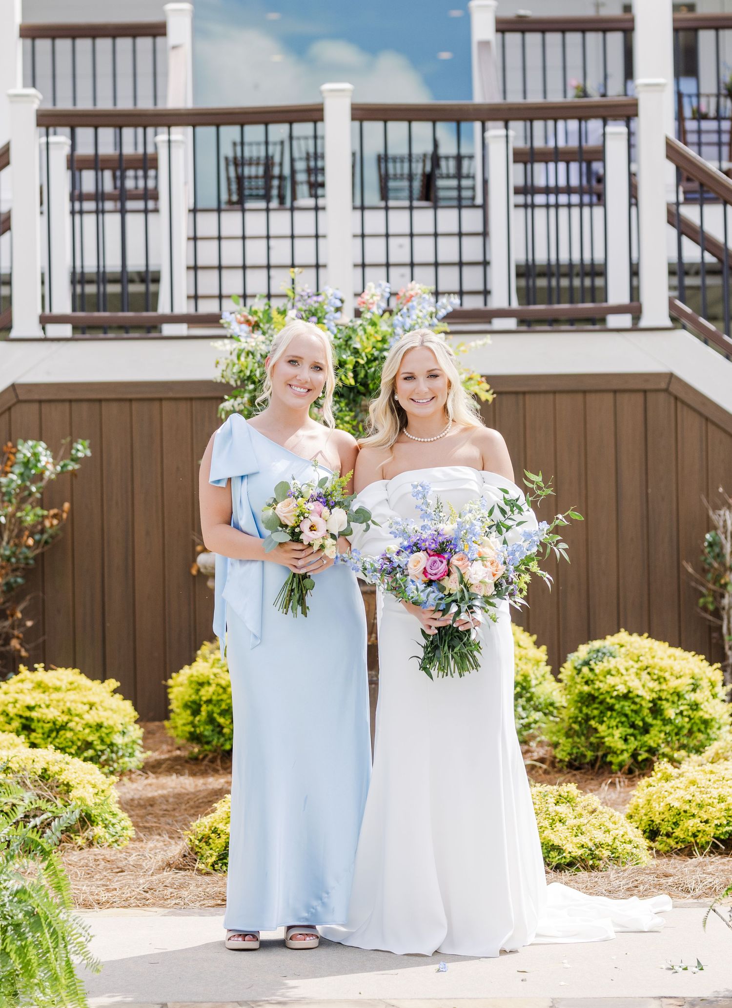 Bride in white strapless gown and bridesmaid in light blue dress hold bouquets outside wedding venue.