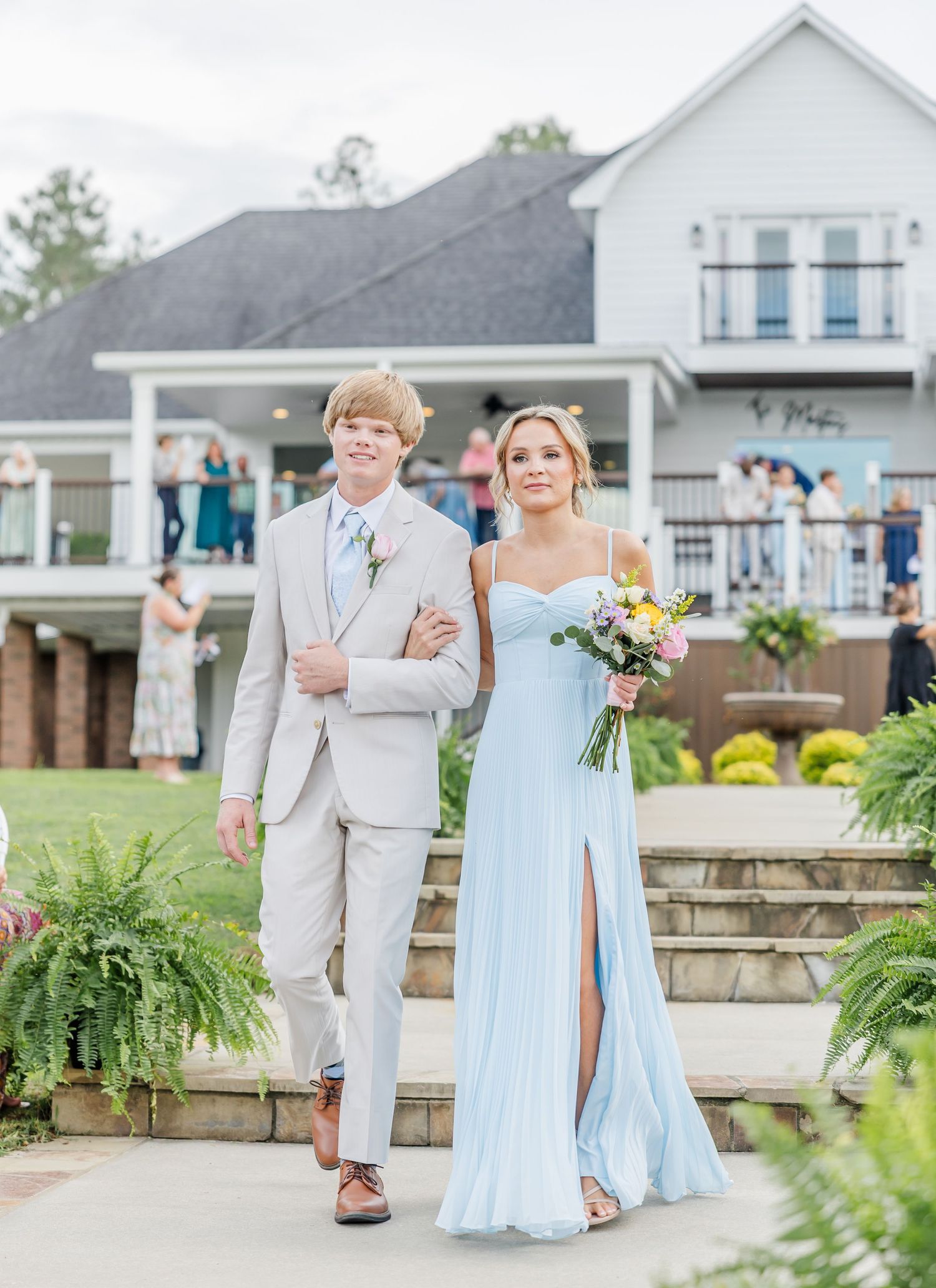 Groomsman in beige suit escorts bridesmaid in light blue gown holding a colorful bouquet at outdoor ceremony.