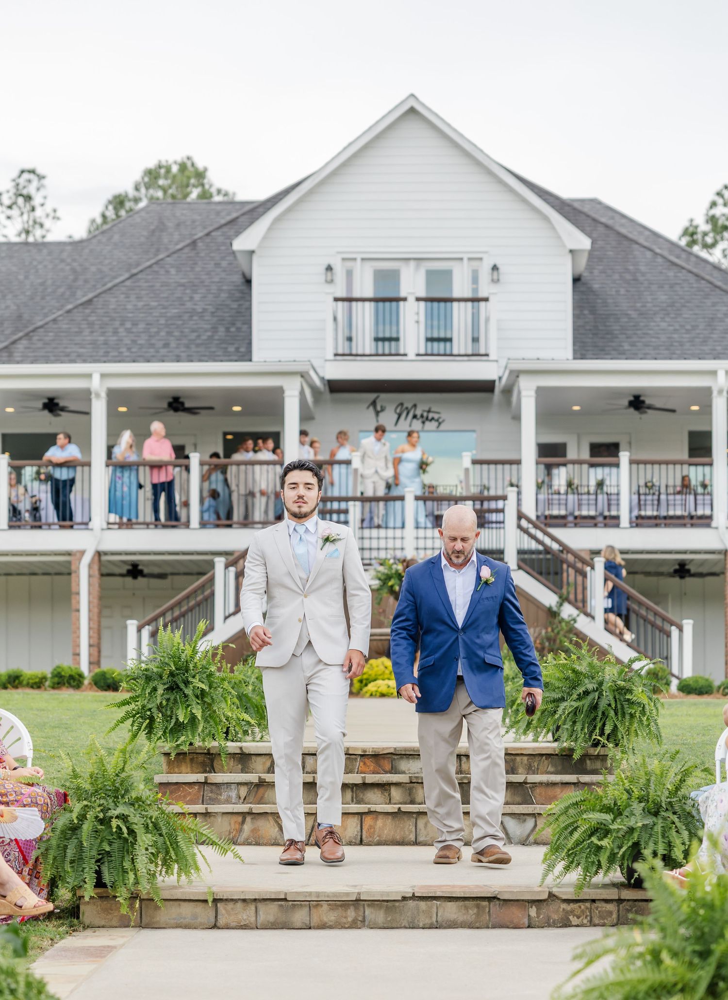 Two grooms walk down outdoor wedding aisle steps before white farmhouse venue with guests on balcony.