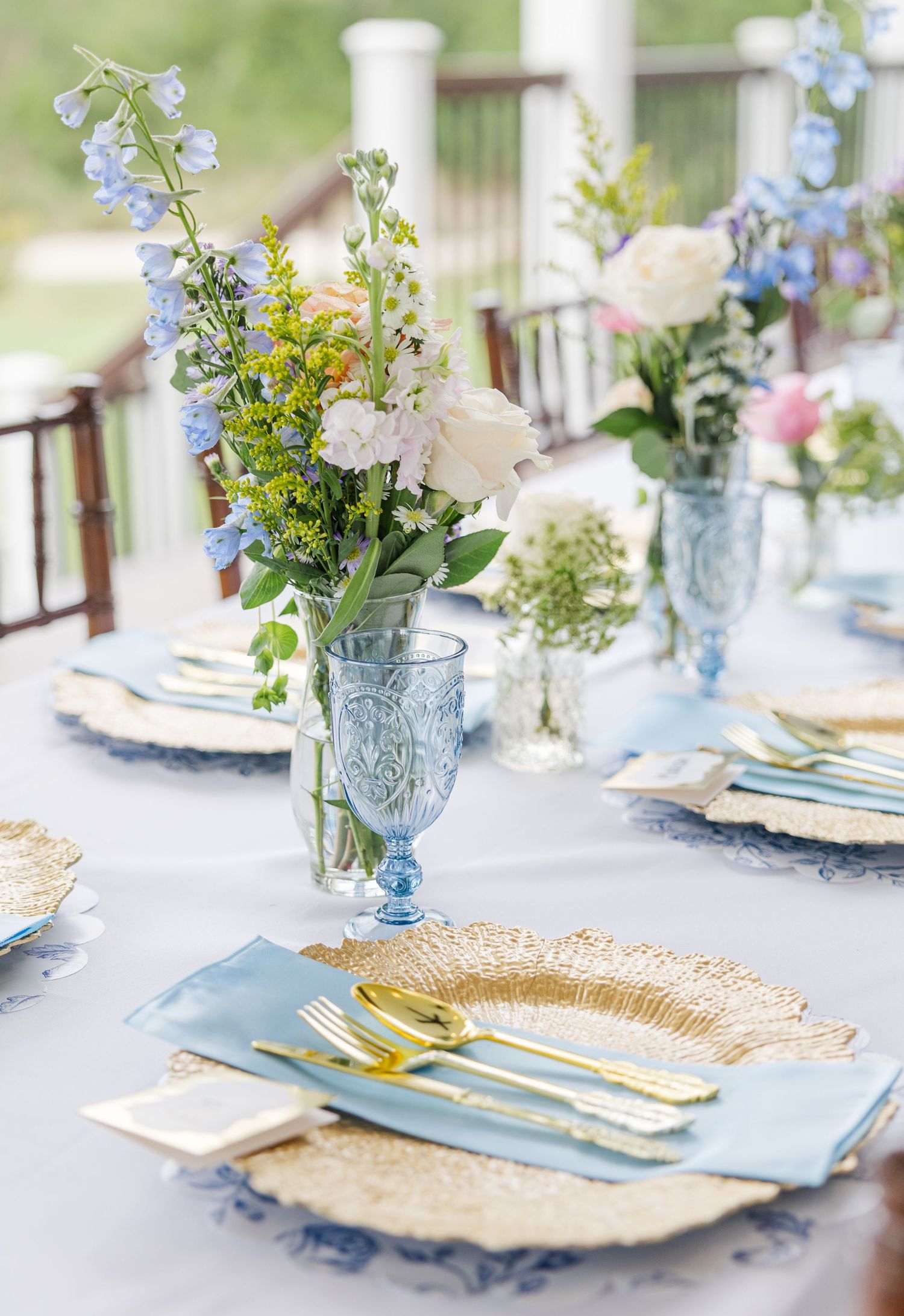 Elegant wedding table setting with blue glassware, gold cutlery, woven chargers, and fresh floral centerpieces.