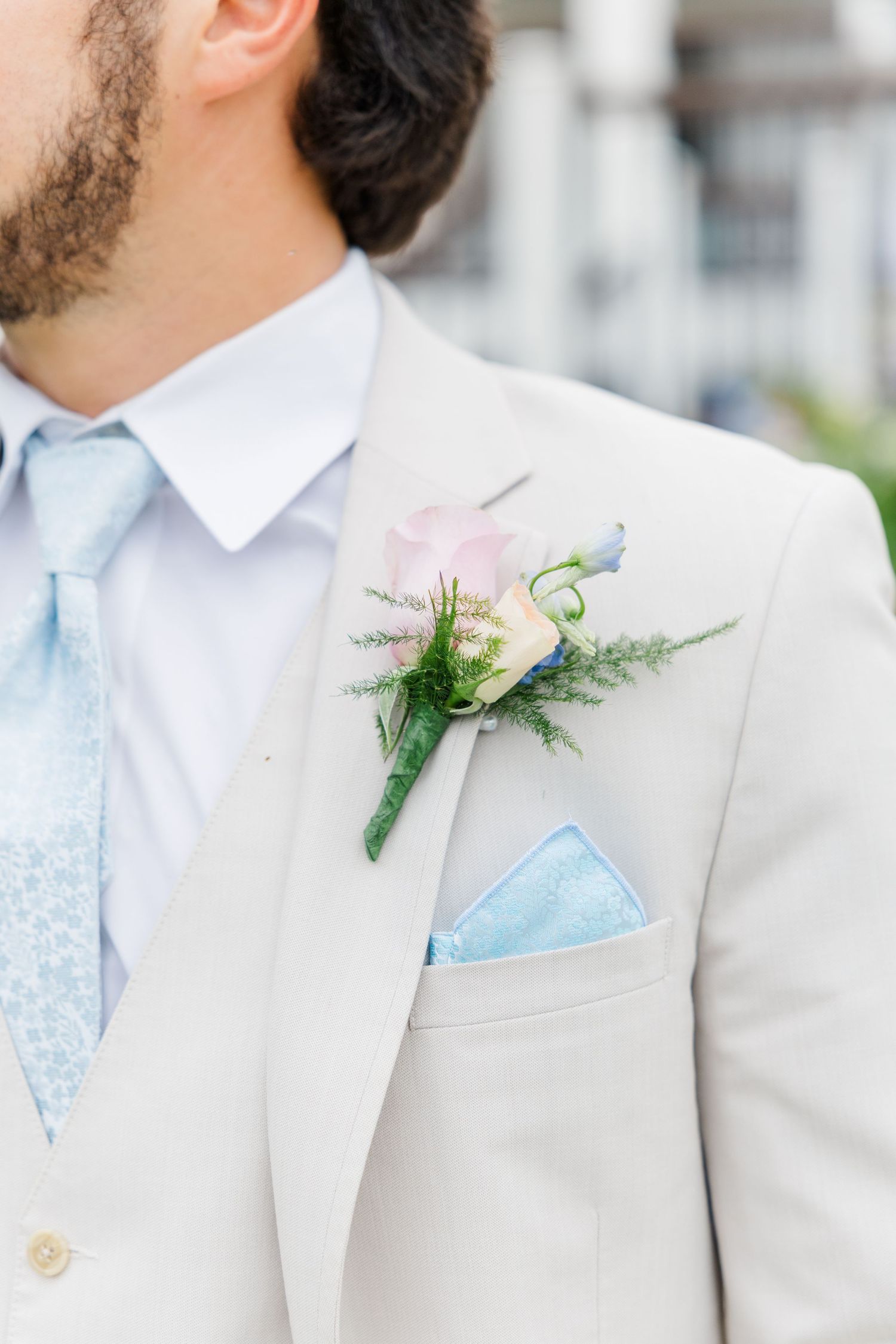 Groom in beige suit with blue tie, floral boutonniere, and light blue pocket square at wedding ceremony.