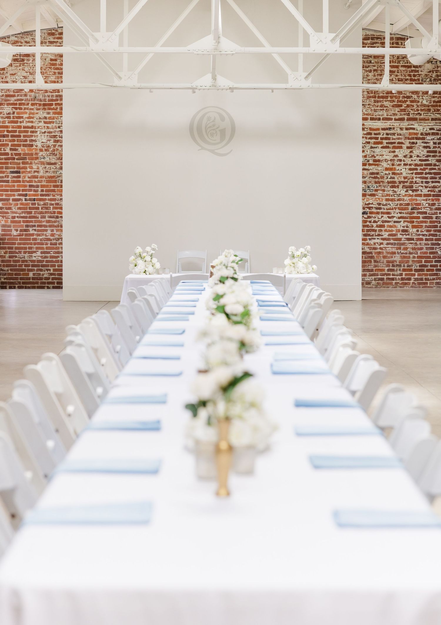 Elegant wedding reception hall with long white tables, blue napkins, white floral centerpieces, and exposed brick walls.