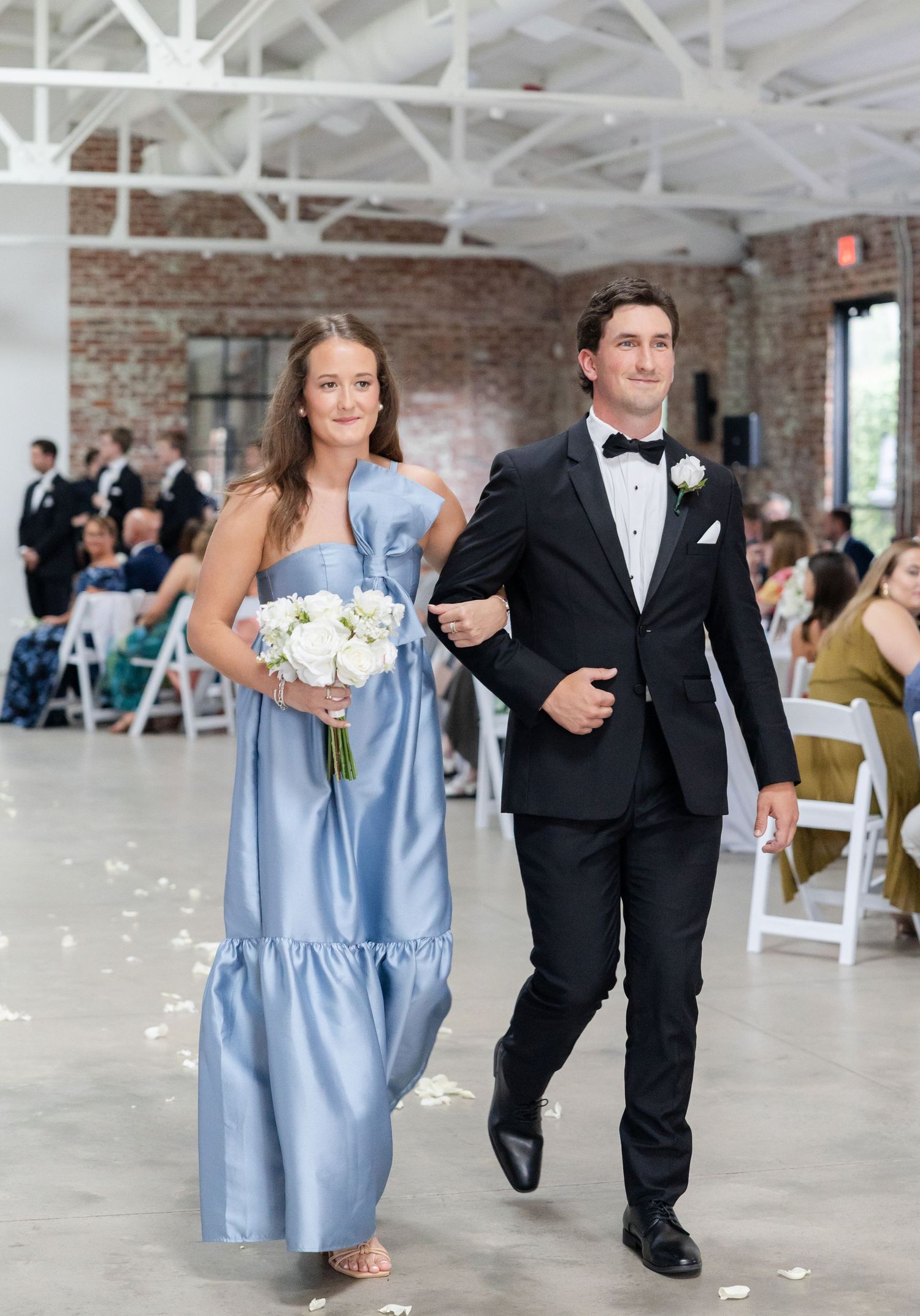 Bridesmaid in blue gown and groomsman in black tuxedo walking down aisle in bright industrial wedding venue.