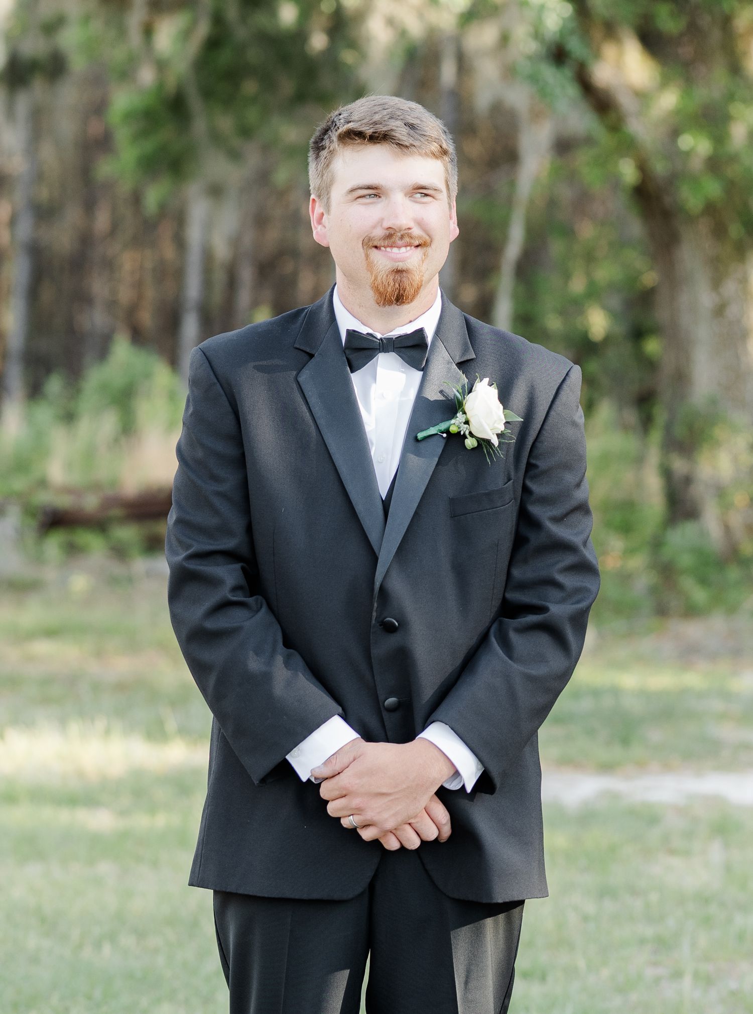Groom in black tuxedo with bow tie and white boutonniere smiling outdoors.