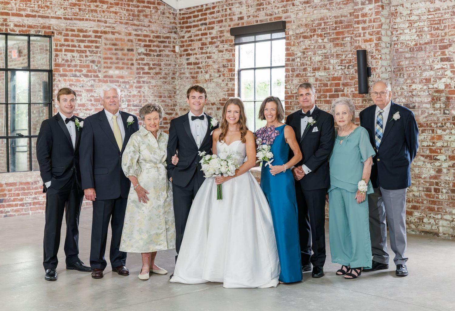 Wedding party family portrait with bride, groom, and extended family posing in front of rustic brick wall venue.