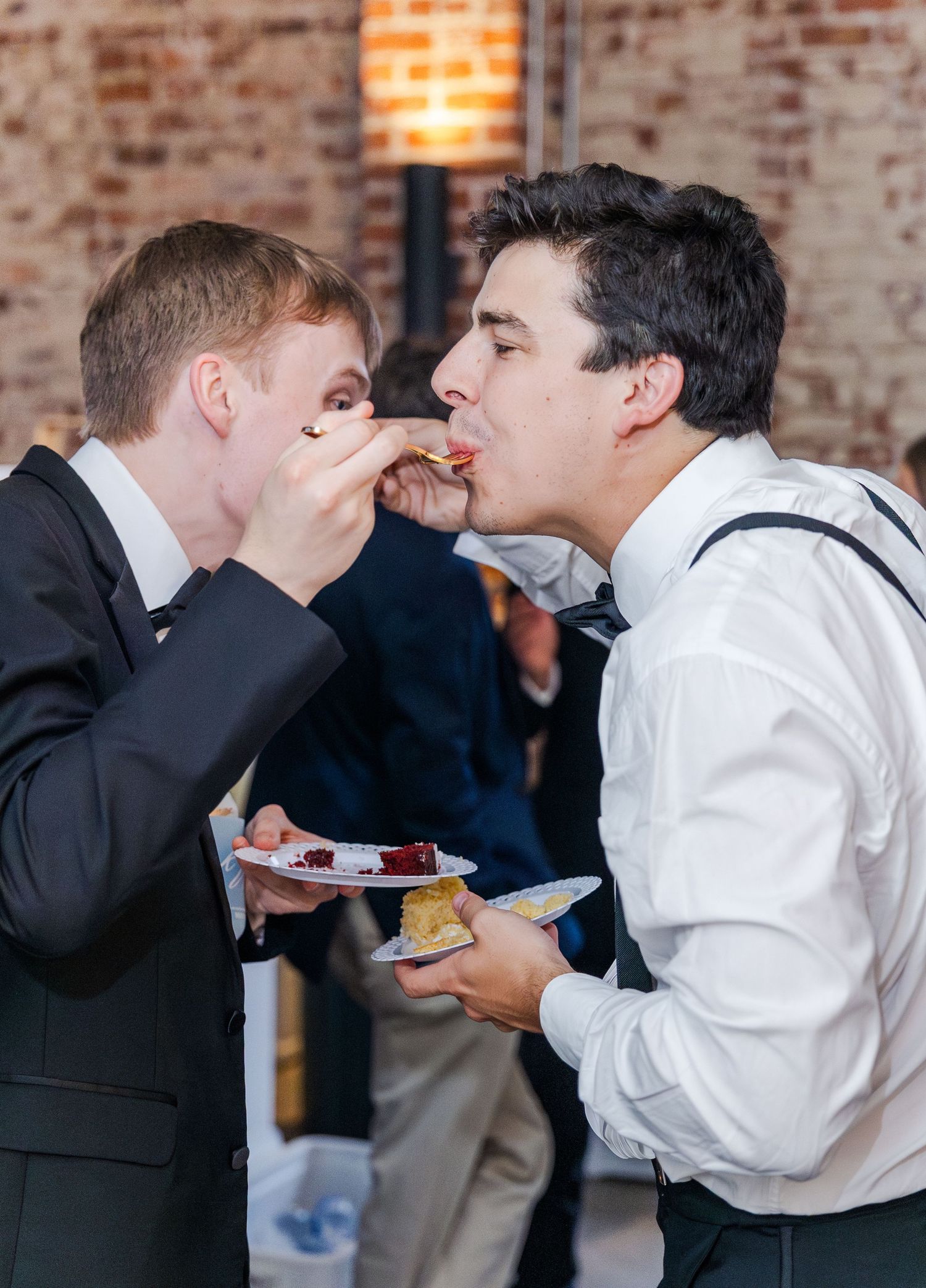 Two young male wedding guests playfully feed each other wedding cake at the reception celebration.