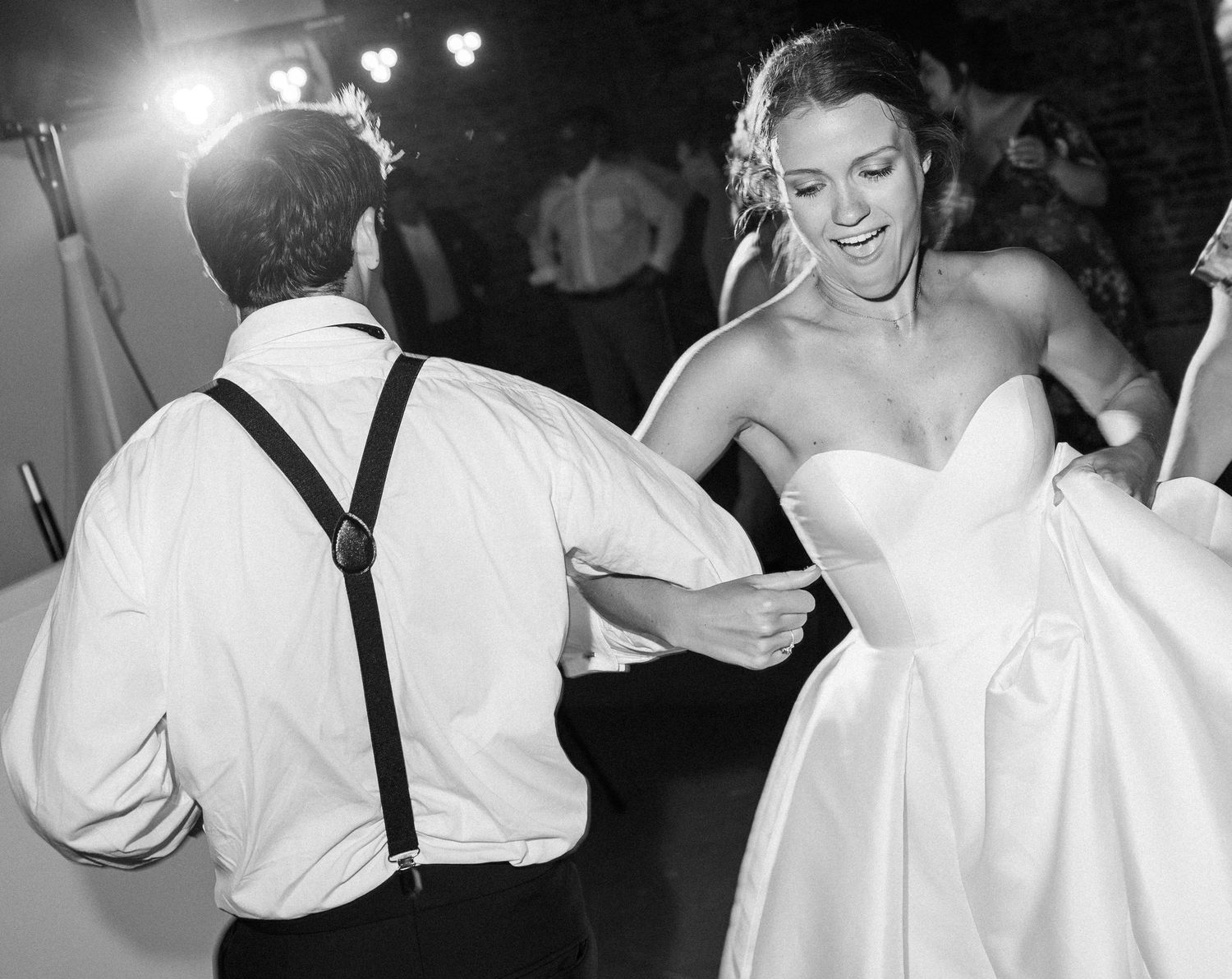 Black and white photo of bride in strapless gown dancing energetically with guest in suspenders at wedding reception.