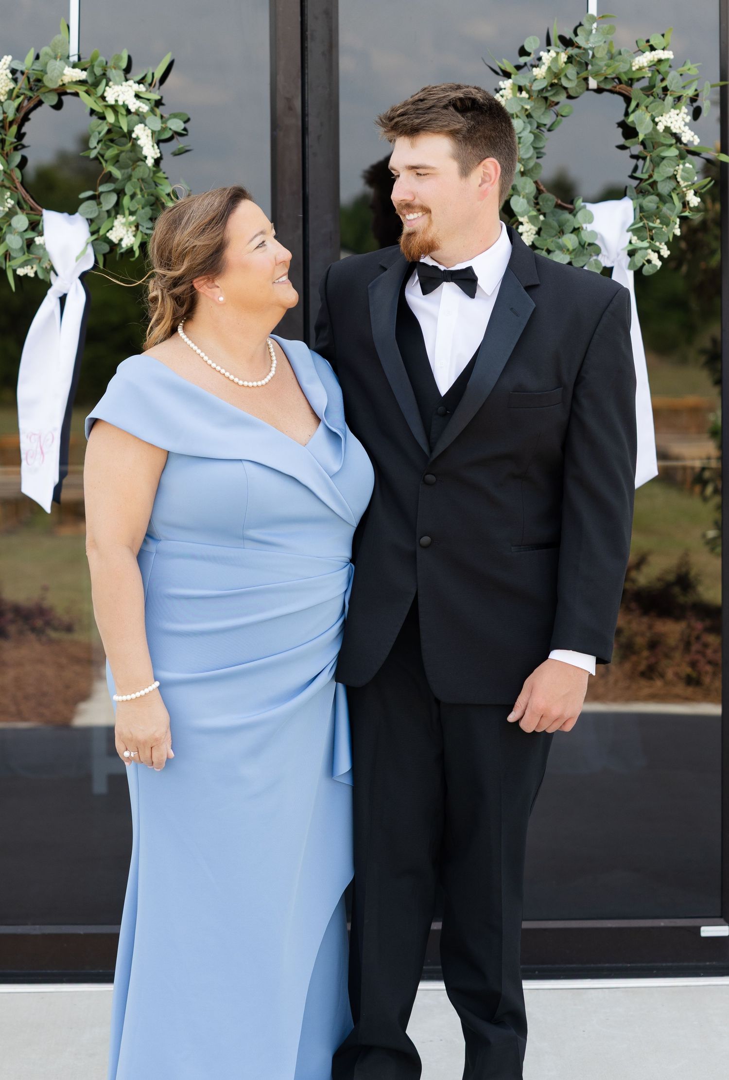 Mother in blue gown and groom in black tuxedo sharing a warm smile outside a wedding venue.