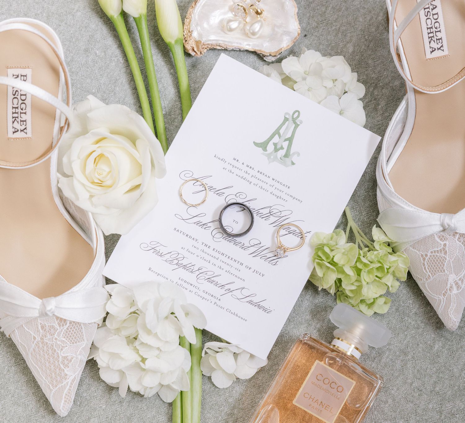 Elegant wedding flat lay with lace heels, rings, invitation, white roses, hydrangeas, and Chanel perfume on gray background.