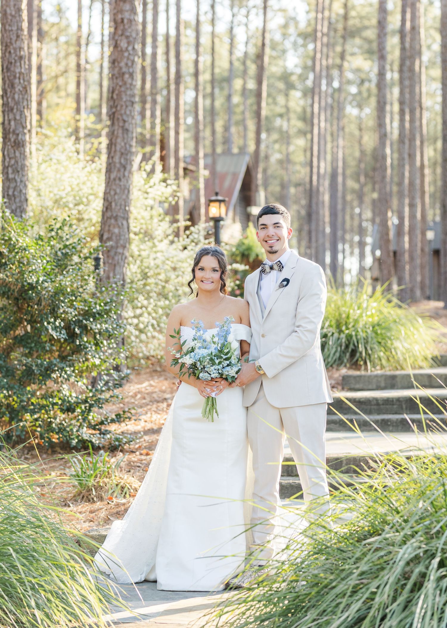 A couple in wedding attire poses together on a wooded path surrounded by tall pine trees and ornamental grasses.