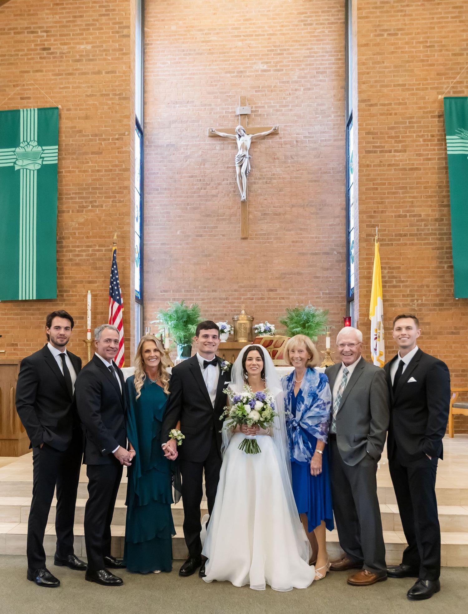 Wedding party poses for formal group photo in church with brick walls and green crosses.