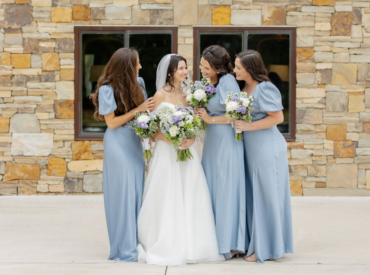 Wedding party in dusty blue dresses and white bridal gown standing outdoors by stone building.