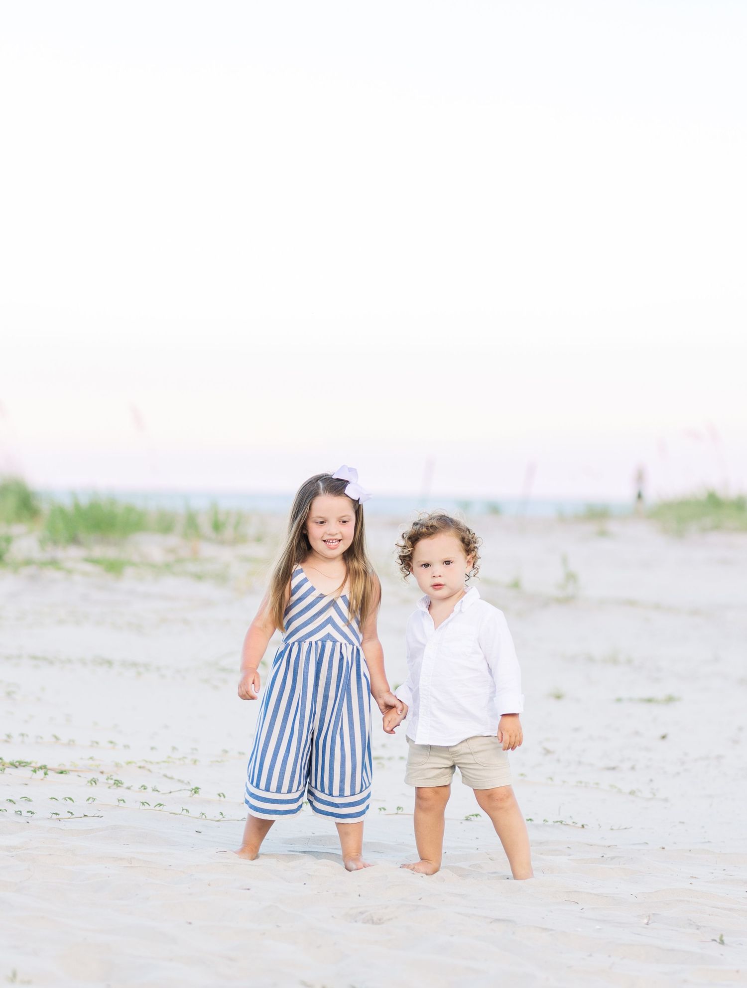 Two young children standing together on a beach, one wearing a striped outfit and one in white.