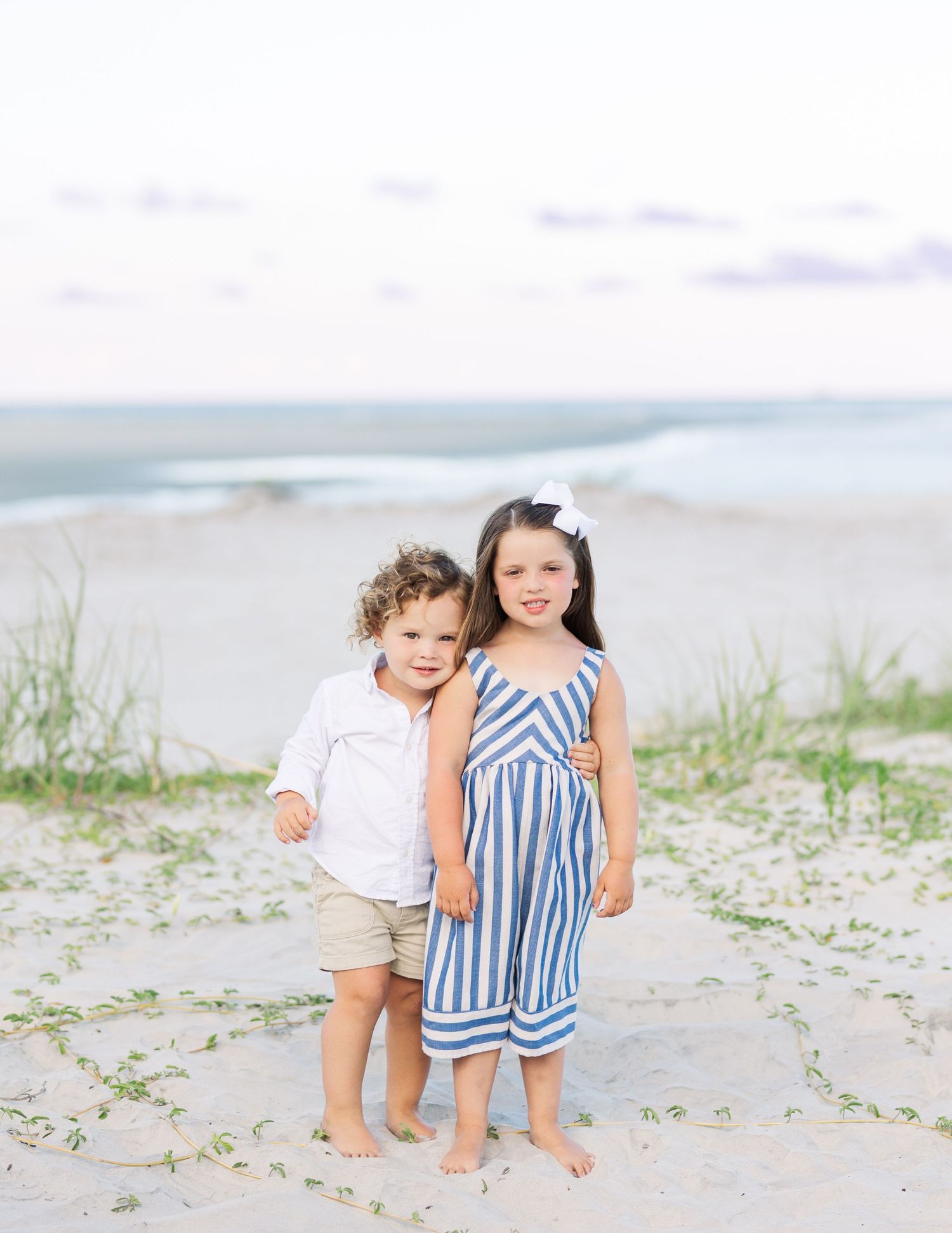 Two small children stand together barefoot on a sandy beach with ocean and grass in the background.