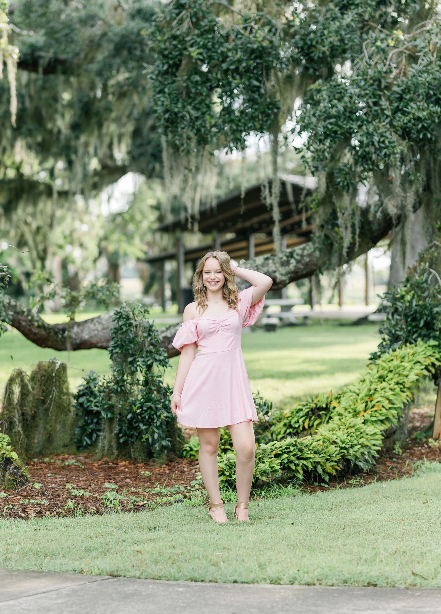 A person in a pink dress poses beneath Spanish moss-draped trees in a lush garden setting.