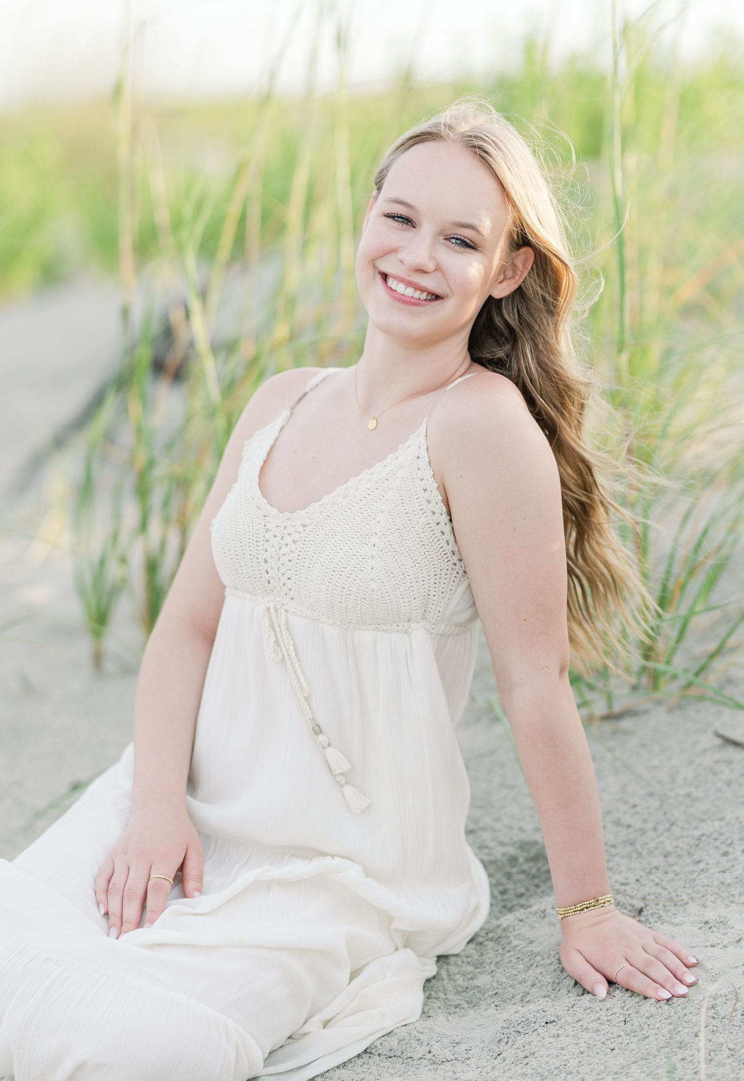 A dreamy beach portrait in a flowing white dress sitting on sandy dunes with sea grass swaying in the breeze.