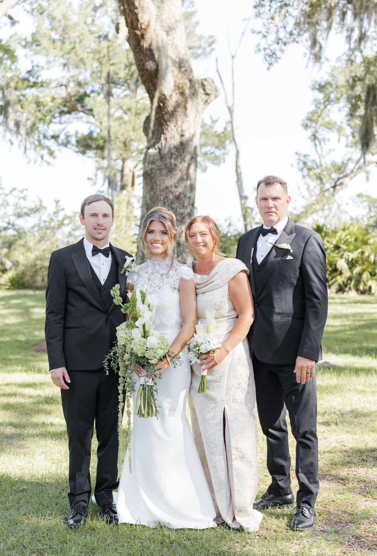 Bride and groom pose with family members outdoors in formal wedding attire, surrounded by lush green trees.