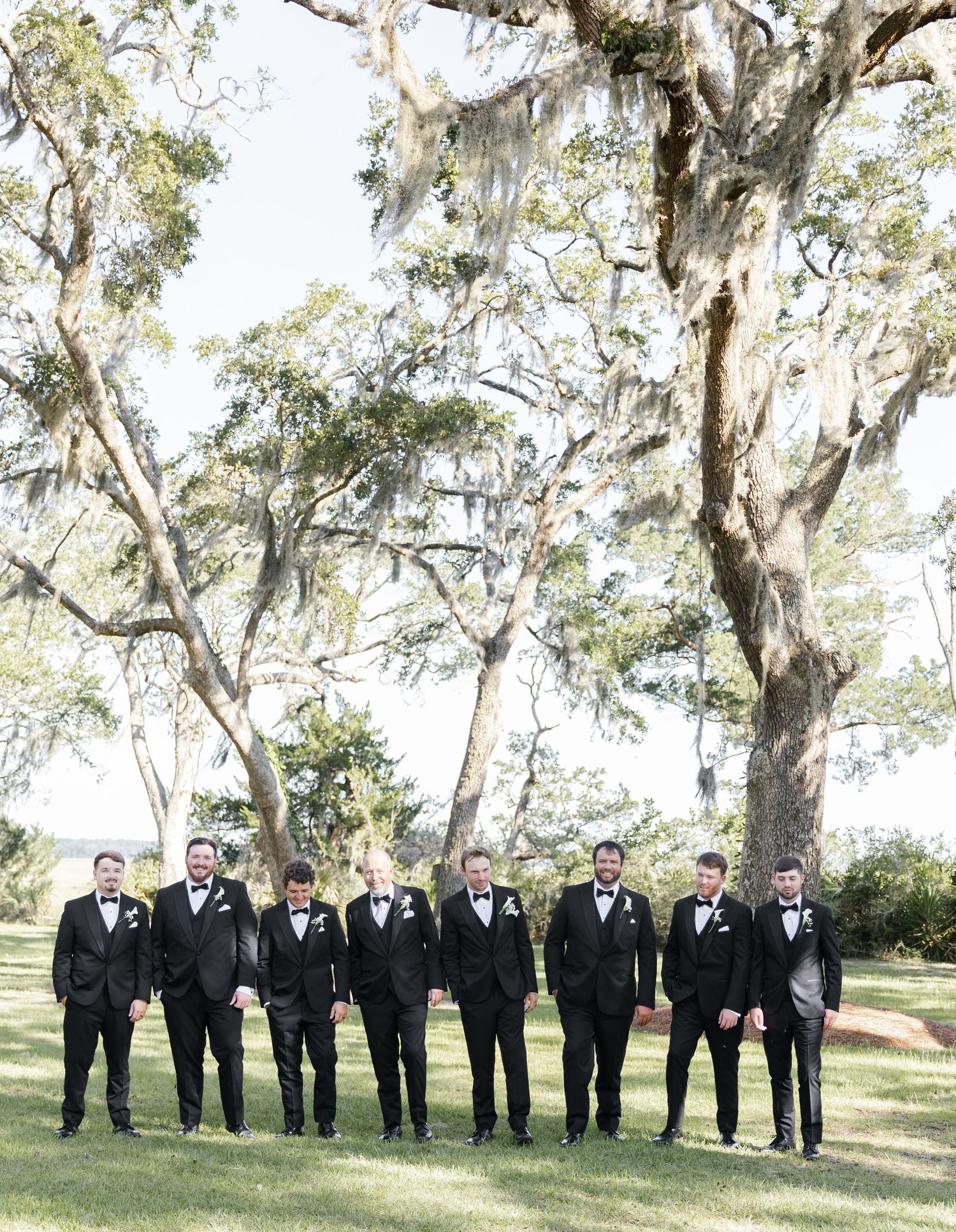 Groom and groomsmen in black tuxedos with bow ties standing outdoors under moss-draped oak trees at a wedding.