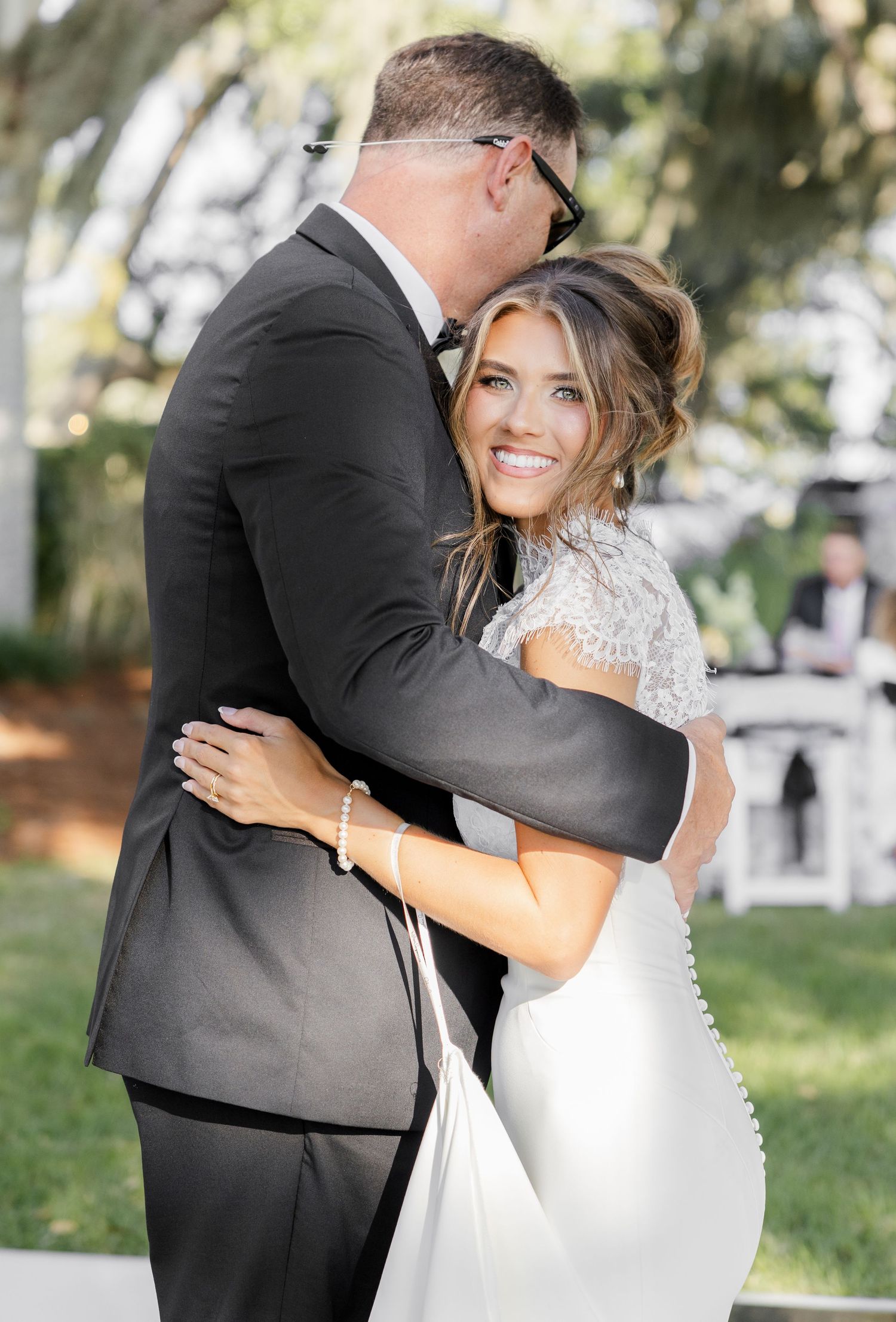 Bride in lace wedding gown smiles while embracing groom in black tuxedo during outdoor wedding ceremony.
