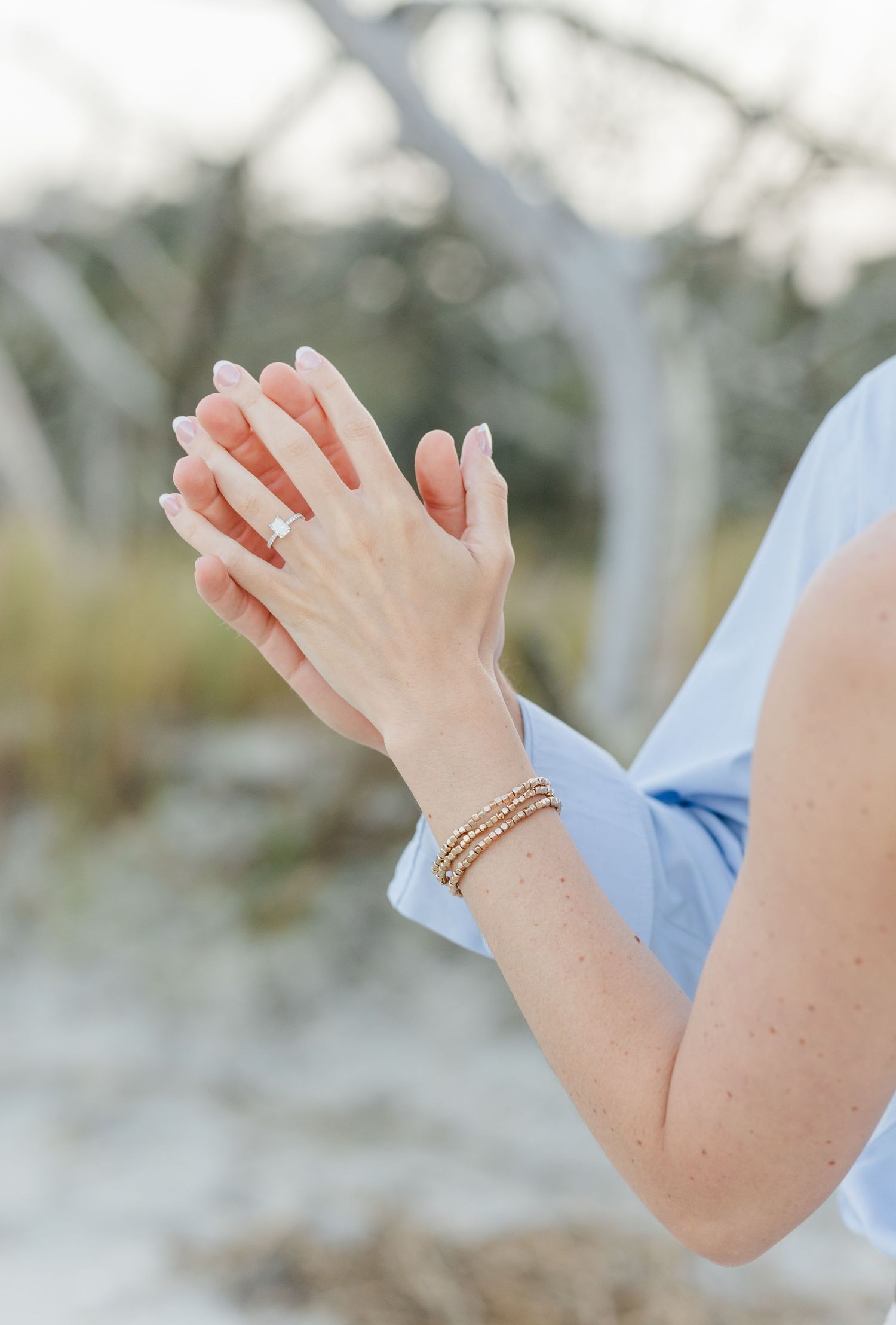 Close-up of a woman's hands displaying an engagement ring and gold bracelets on a beach.
