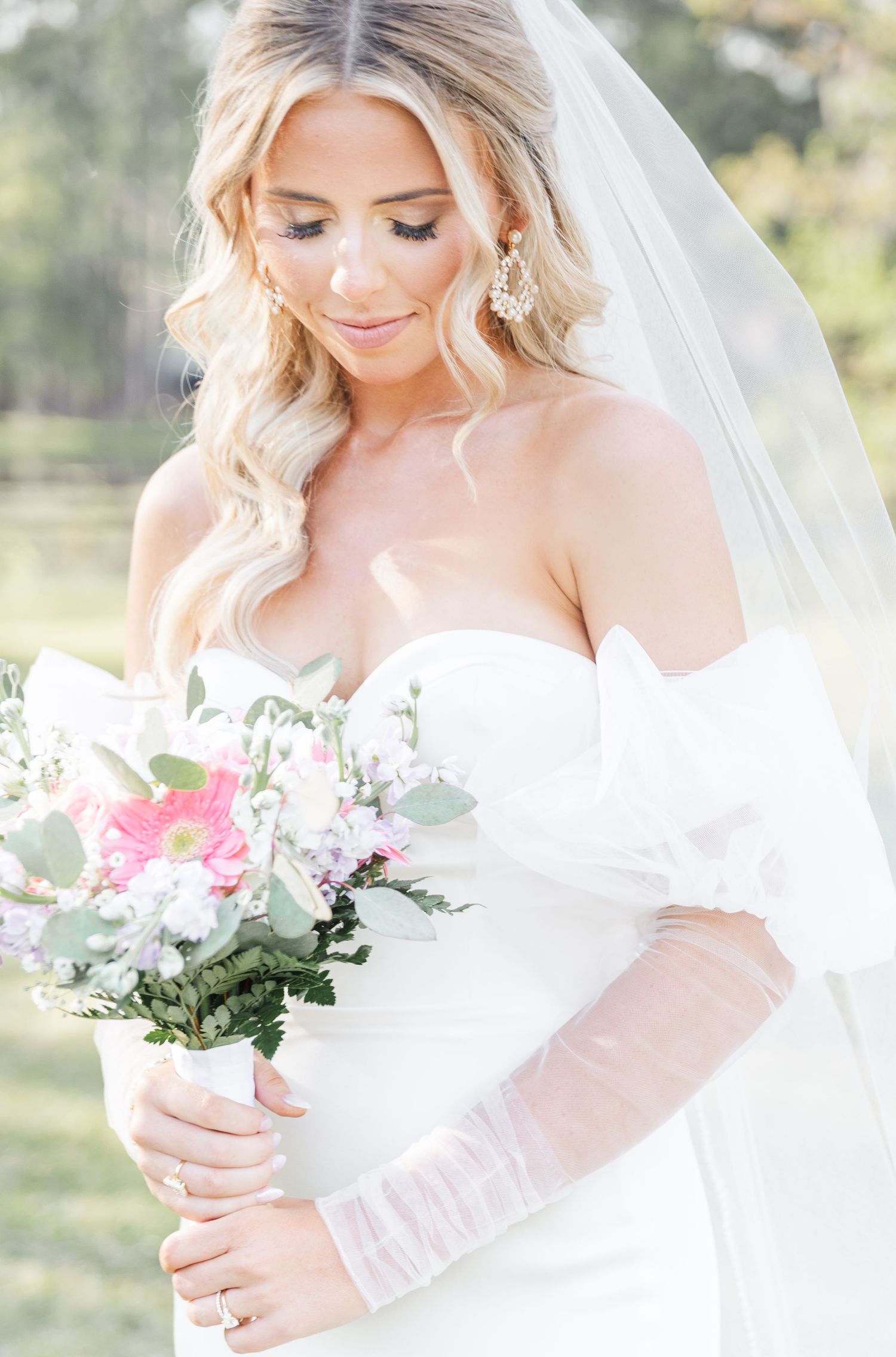 Close-up of bride holding pink and white bouquet with delicate lace veil and wavy hair.