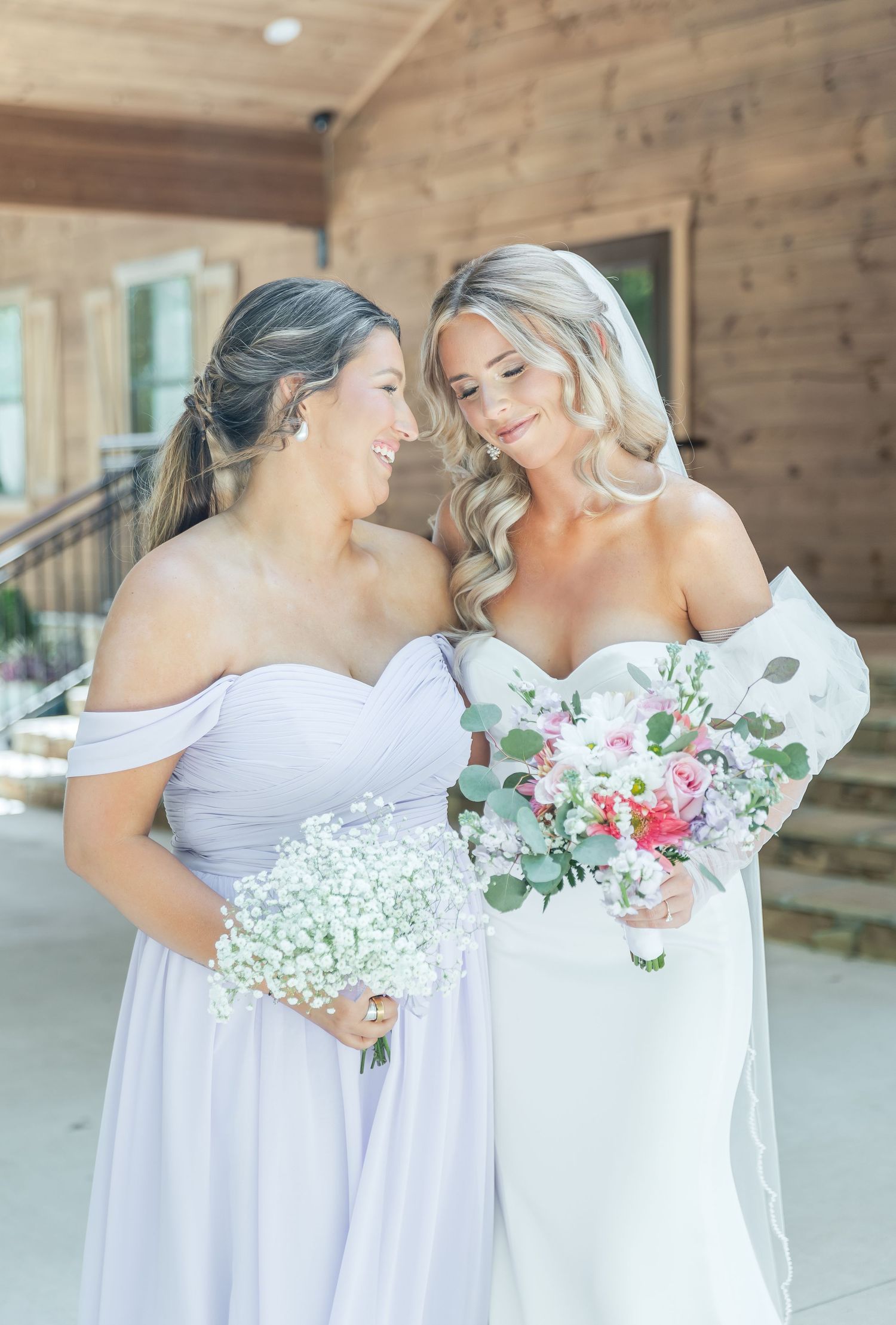 Two women in bridal party attire share laughter while holding bouquets against wooden backdrop.
