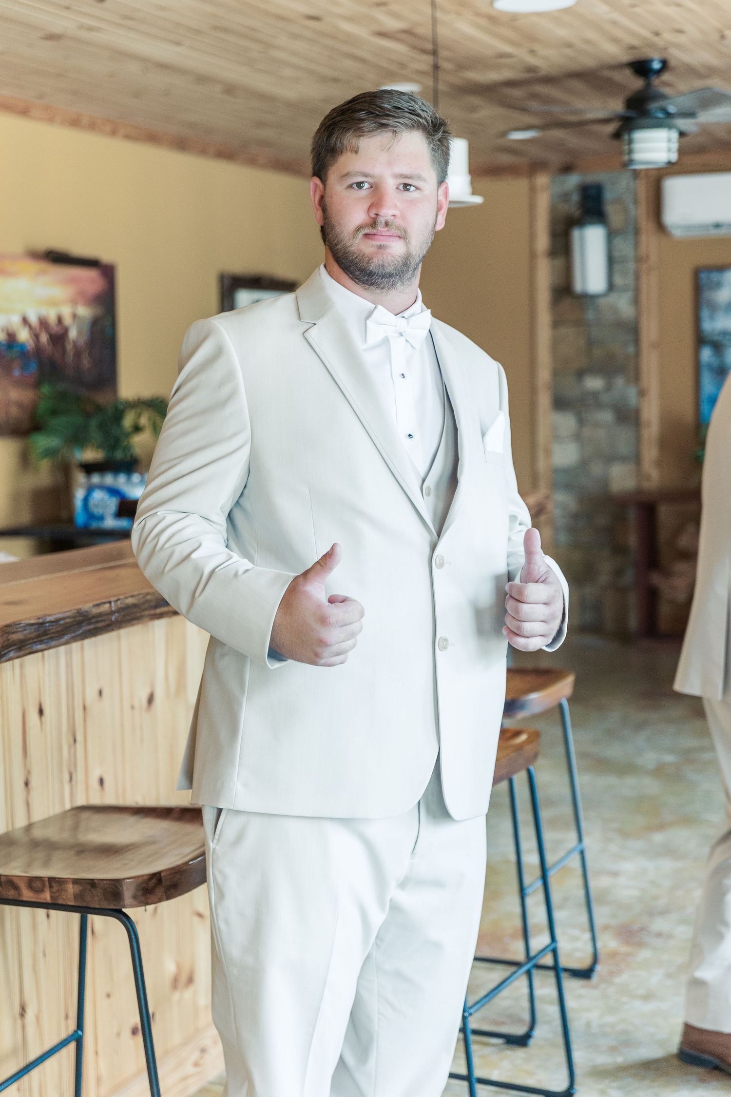 Groom in an all-white tuxedo gives thumbs up while standing in rustic venue.