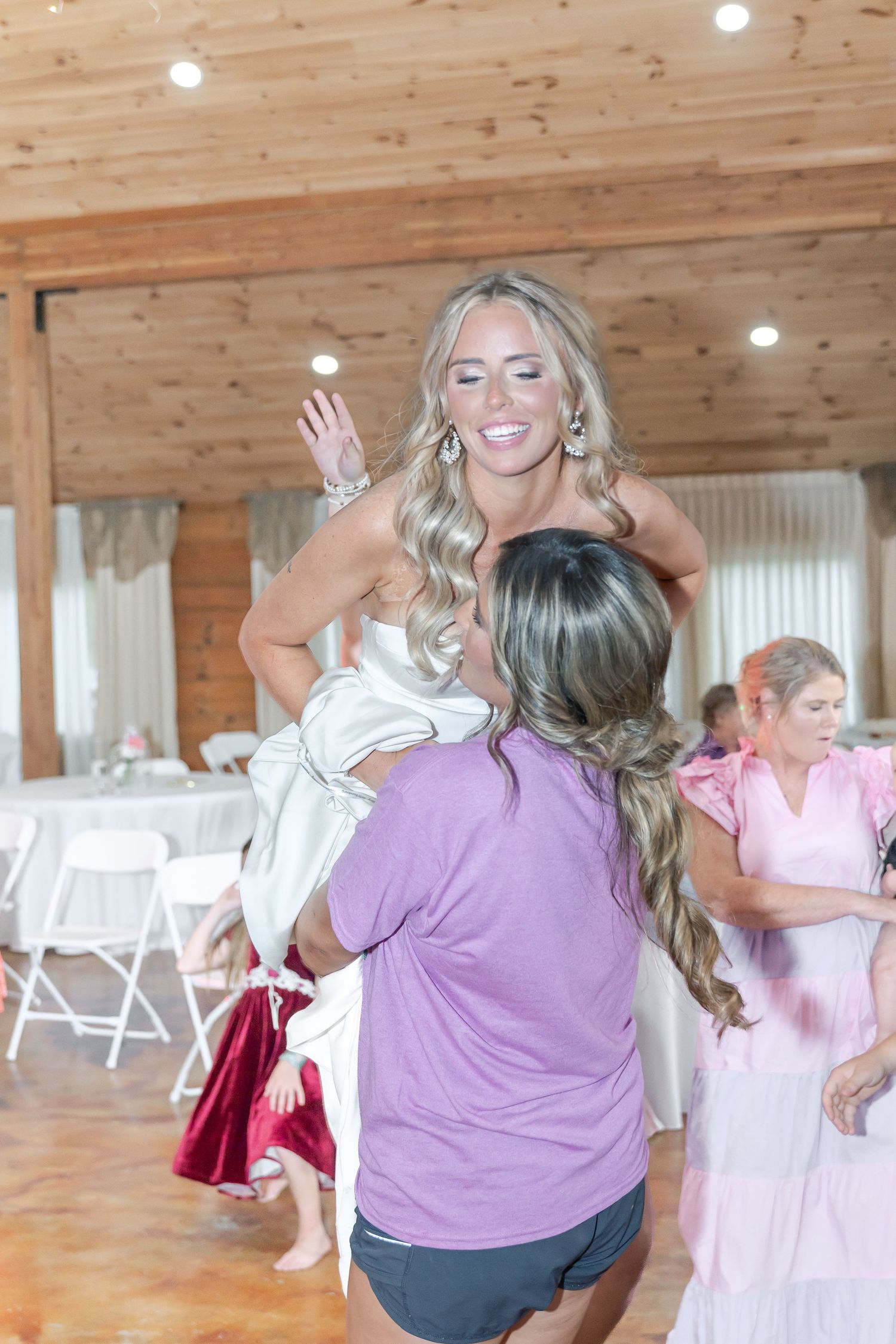 Dancers celebrating at a rustic wedding reception with wooden walls and white folding chairs.