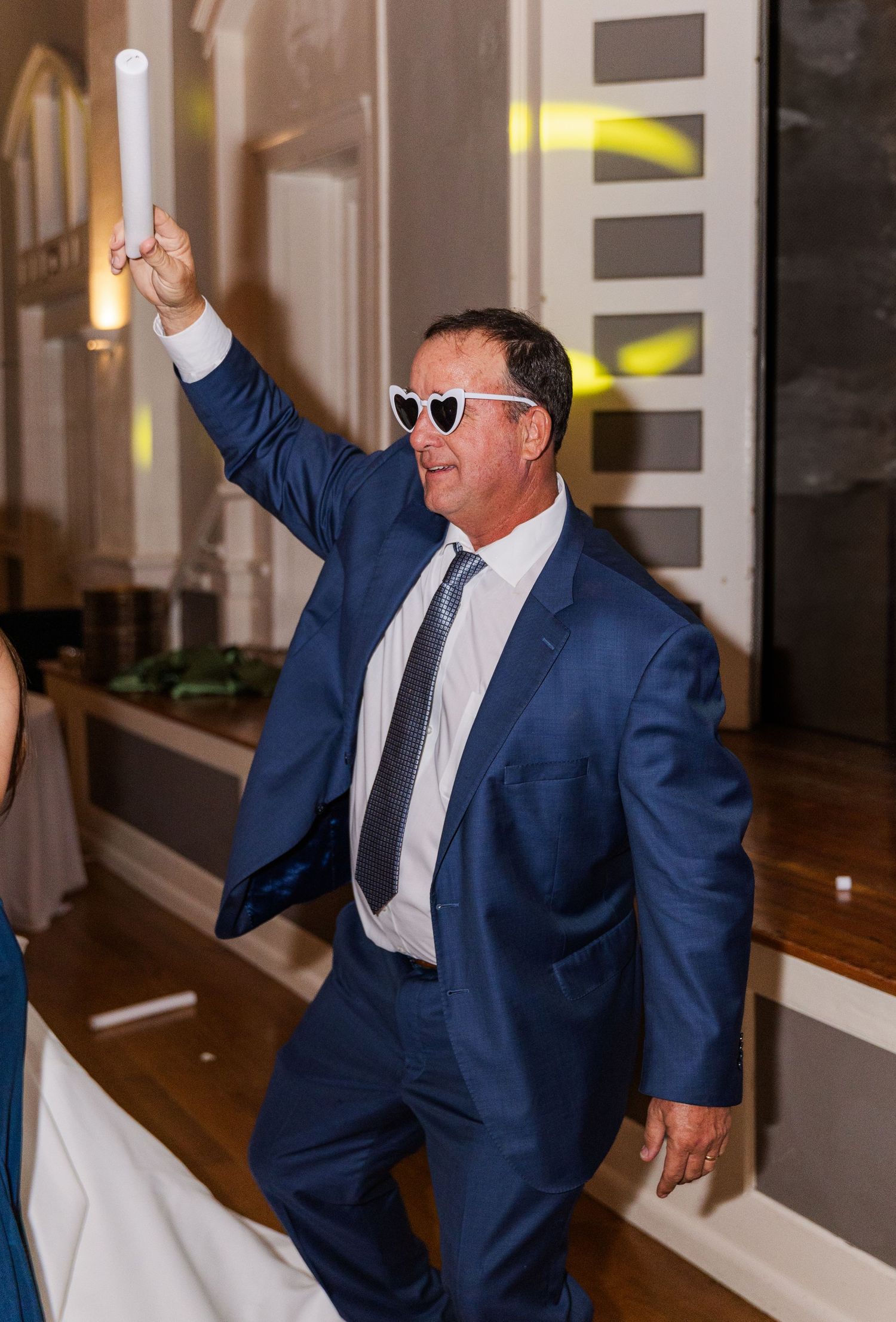 Man in blue suit raises glow stick wearing white heart sunglasses while dancing at wedding reception.