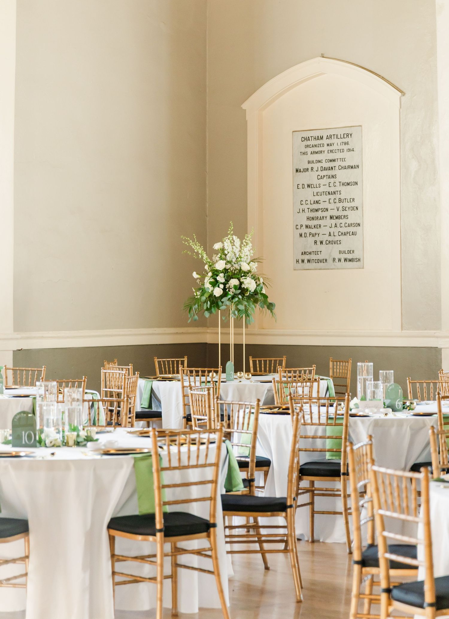 Elegant wedding reception hall with gold chiavari chairs, white tablecloths, sage green accents, and tall floral centerpieces.