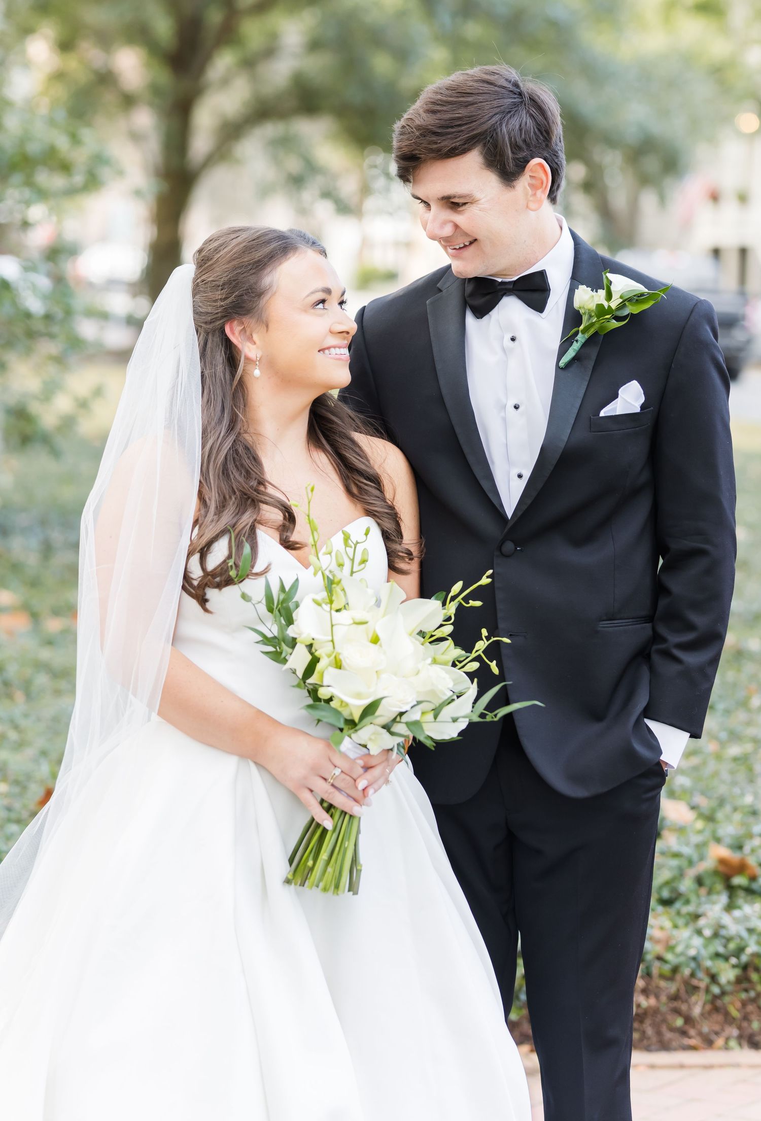 Smiling bride and groom share a tender moment, bride holding a white floral bouquet outdoors.