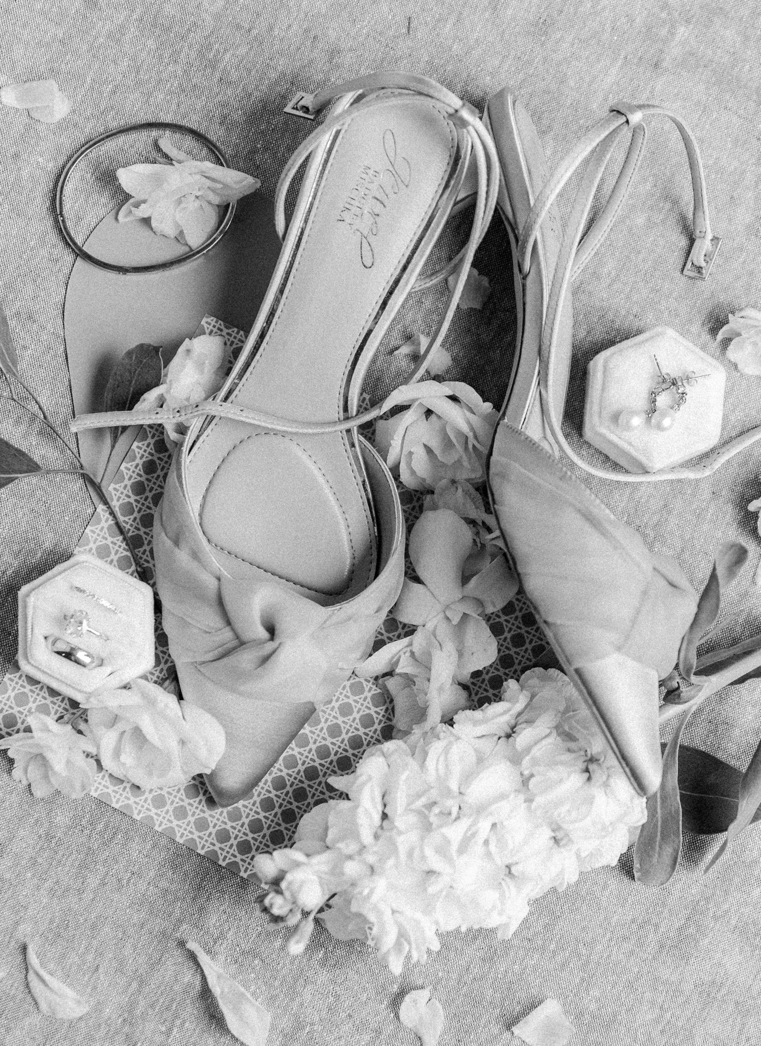 Black and white flat lay of bridal heels, floral accessories, and jewelry boxes arranged elegantly on a patterned surface.