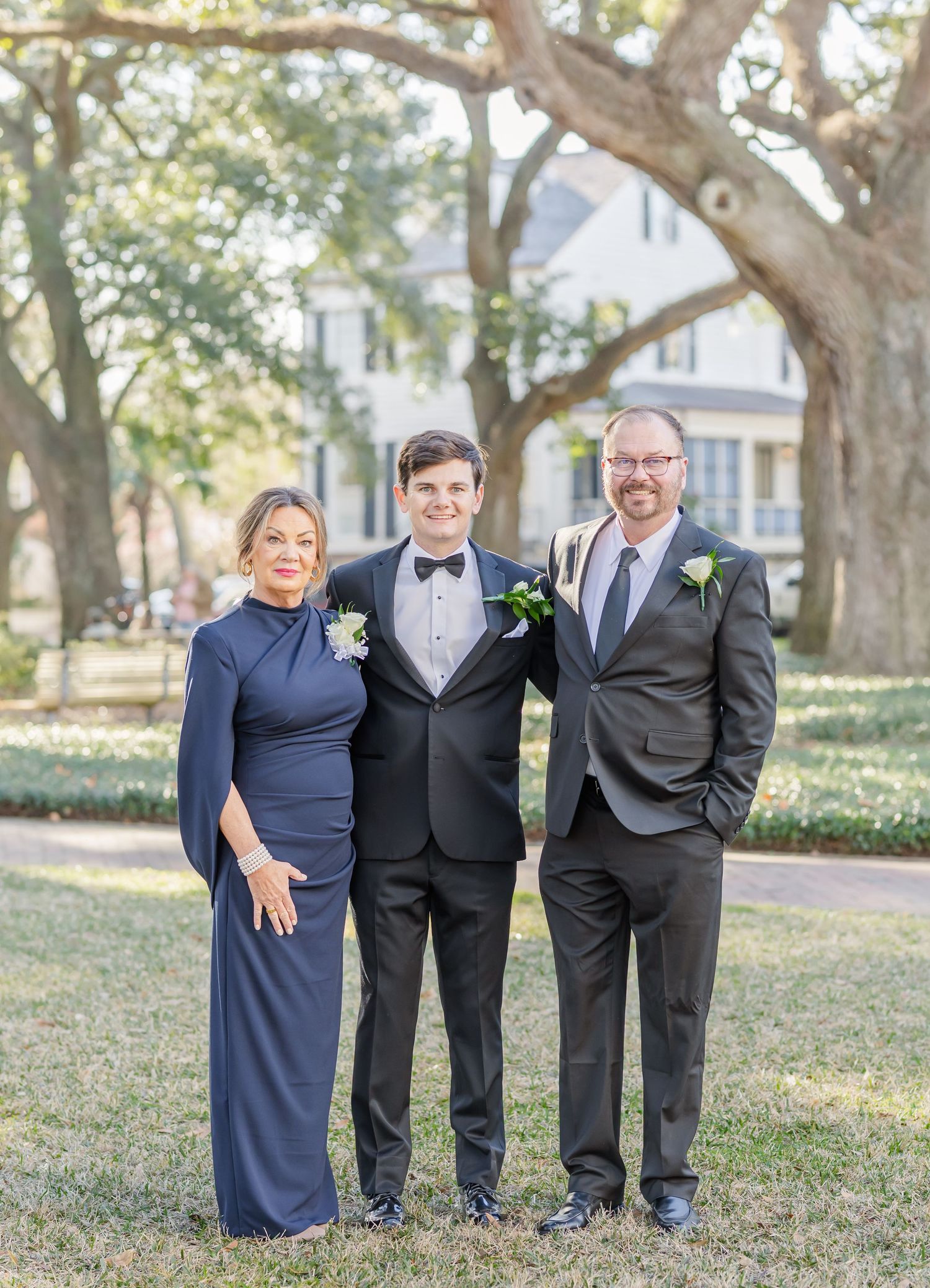 Groom in tuxedo poses with parents in formal attire outdoors under oak trees at a wedding celebration.