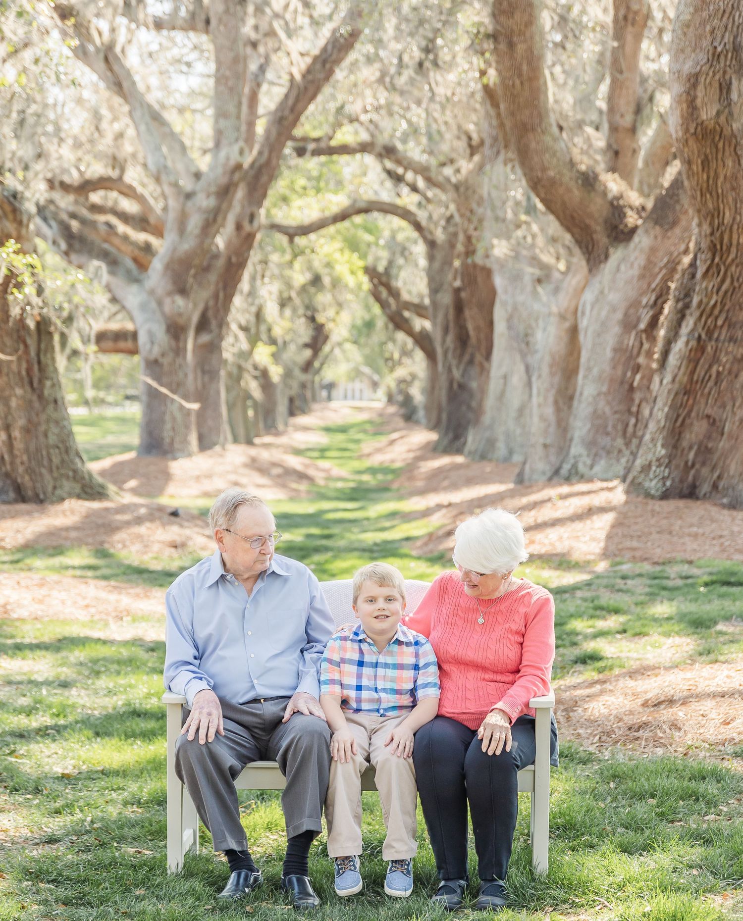 Three family members seated on white bench along scenic oak tree path.