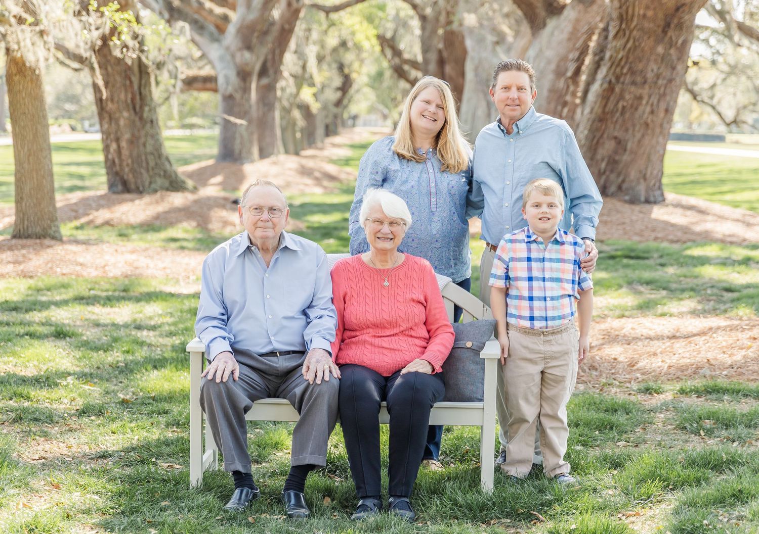 Multi-generational family portrait on bench under large oak trees.